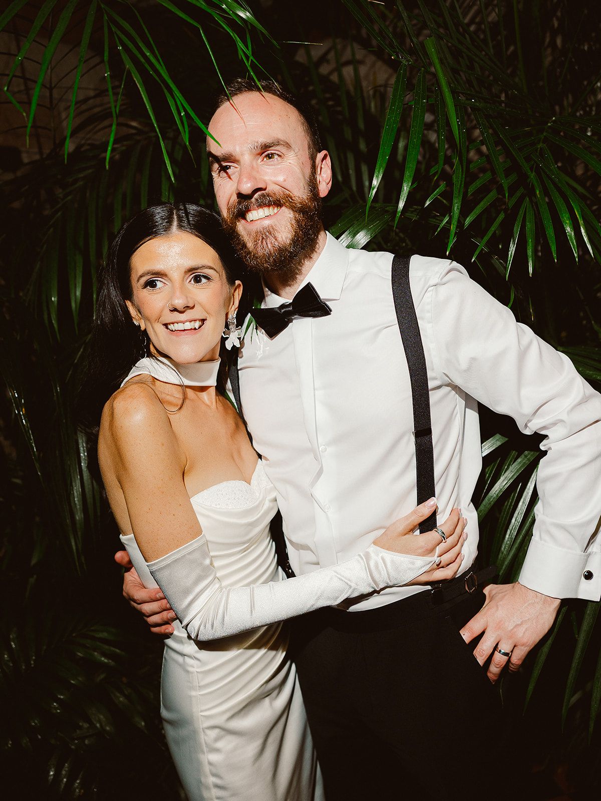 A couple in wedding attire share playful moments during their reception in a dark tropical setting with palm fronds.