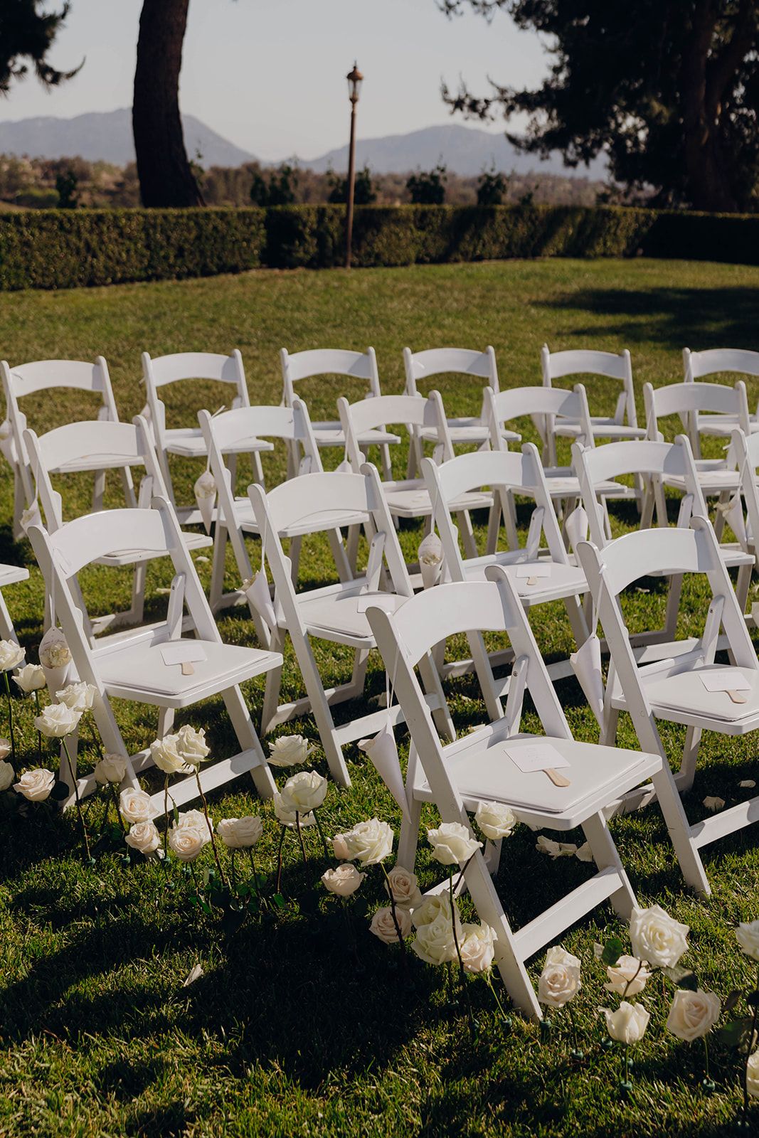 White folding chairs arranged for an outdoor wedding ceremony with white floral decorations along the aisle on a manicured lawn.