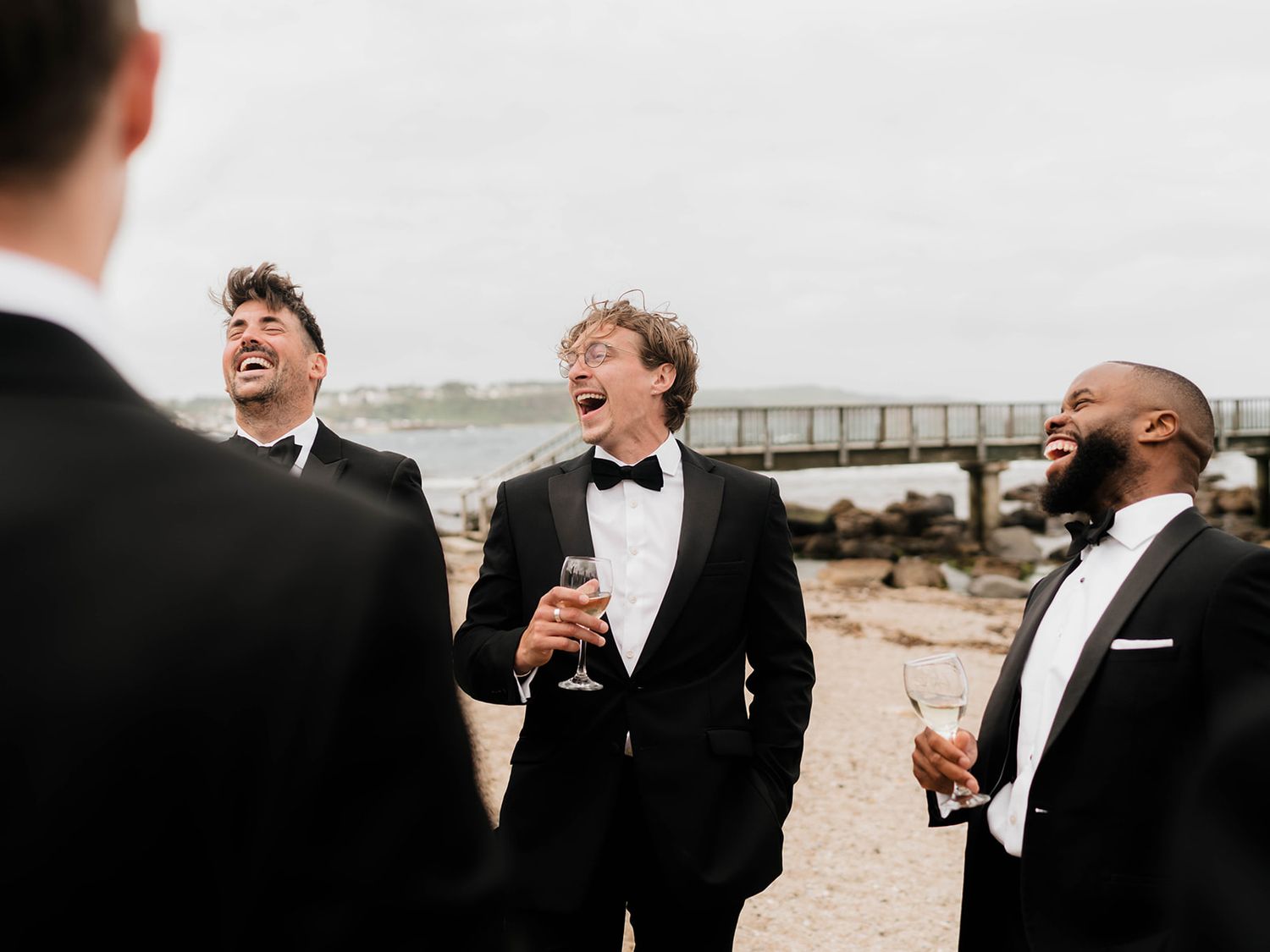 Groomsmen sharing a joyful laugh together while holding drinks on a beach during a wedding celebration.