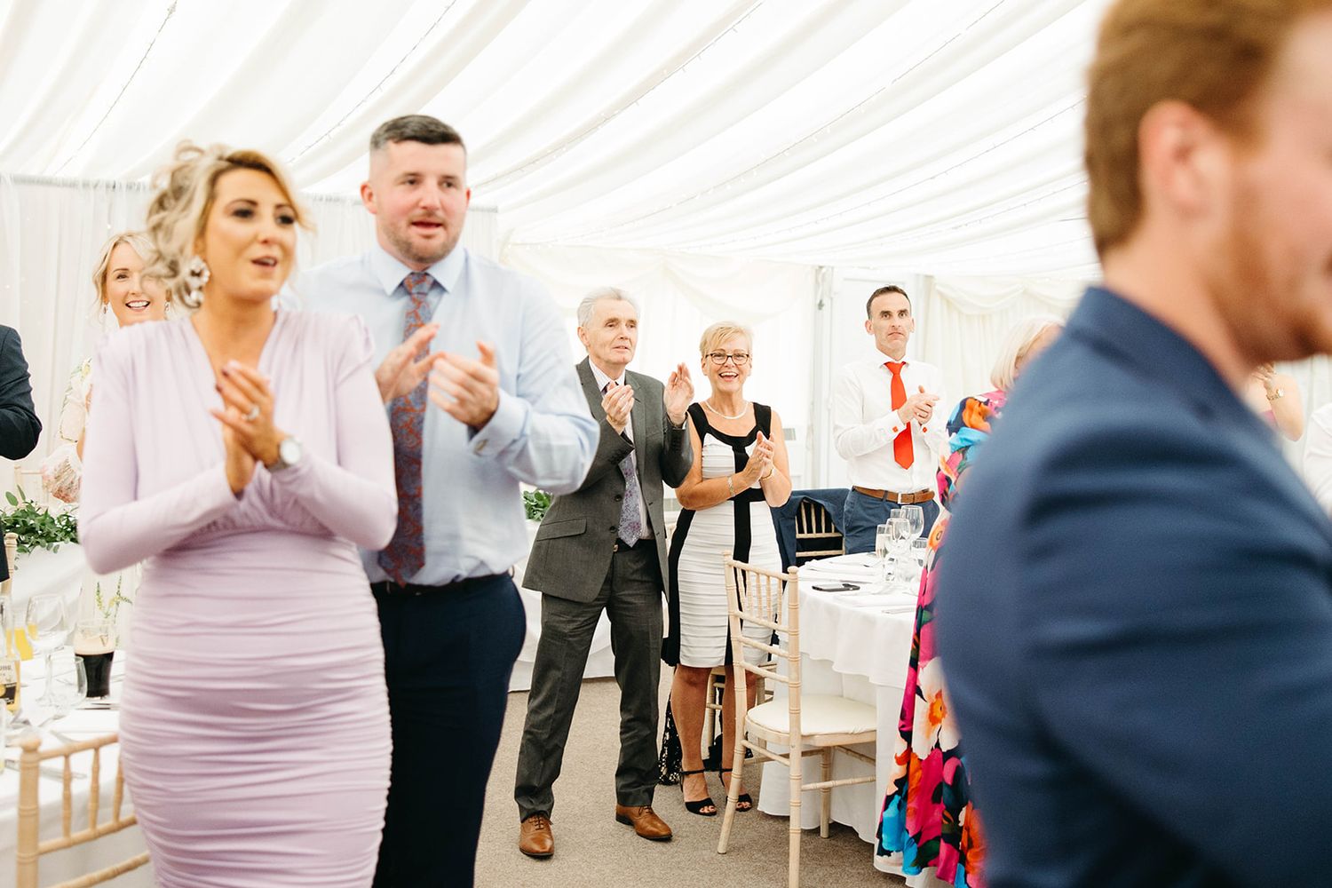 Wedding guests standing in a white tent venue with emotional expressions during a reception celebration.