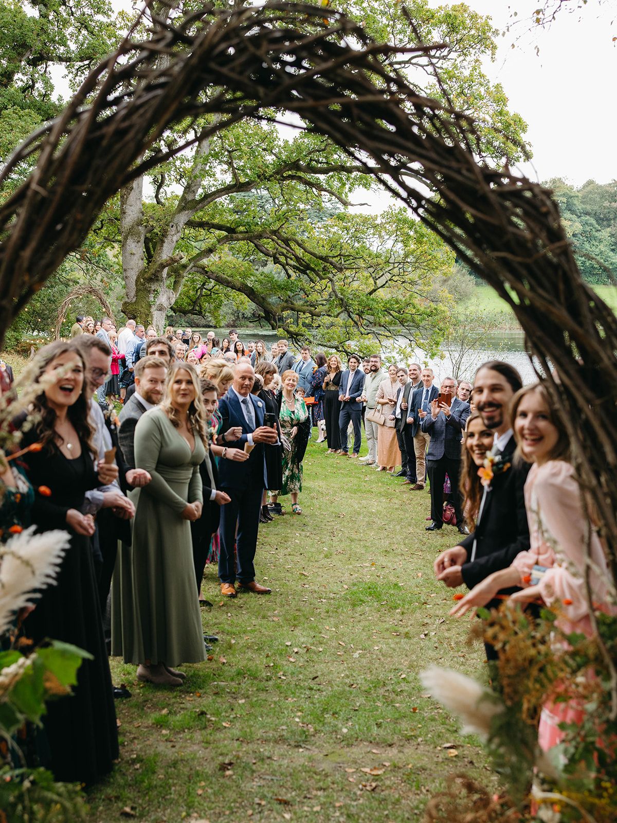 Wedding guests line up under a rustic wooden arch made of twisted branches in an outdoor garden ceremony.