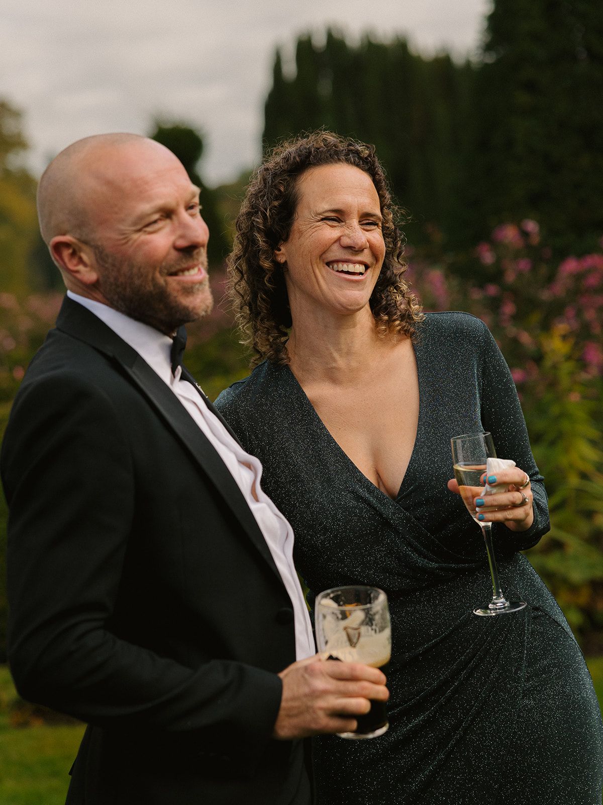 A couple shares a joyful moment together at an outdoor event, holding wine glasses and dressed in formal attire.
