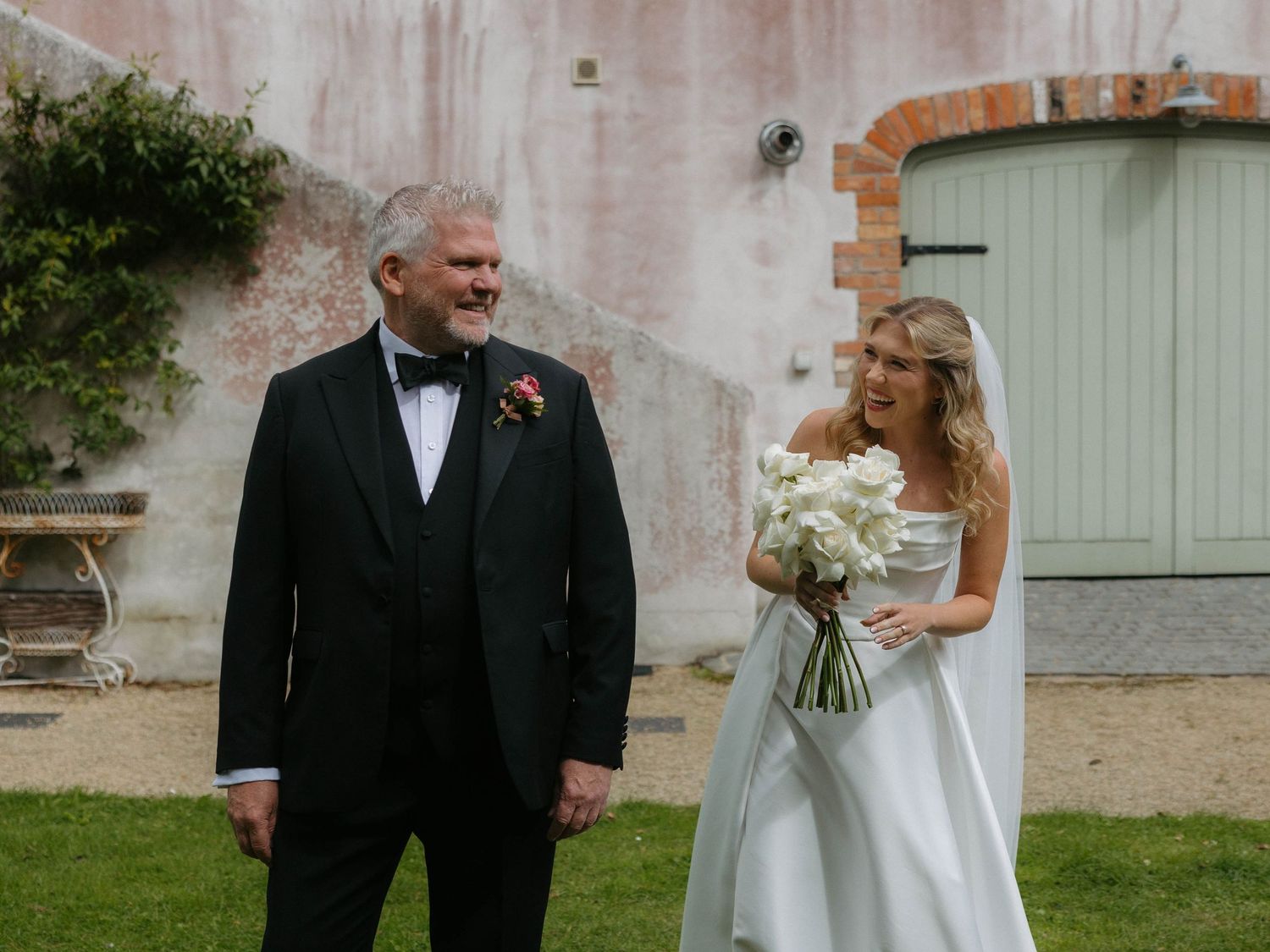 Bride and father share emotional moment before ceremony against pink wall.