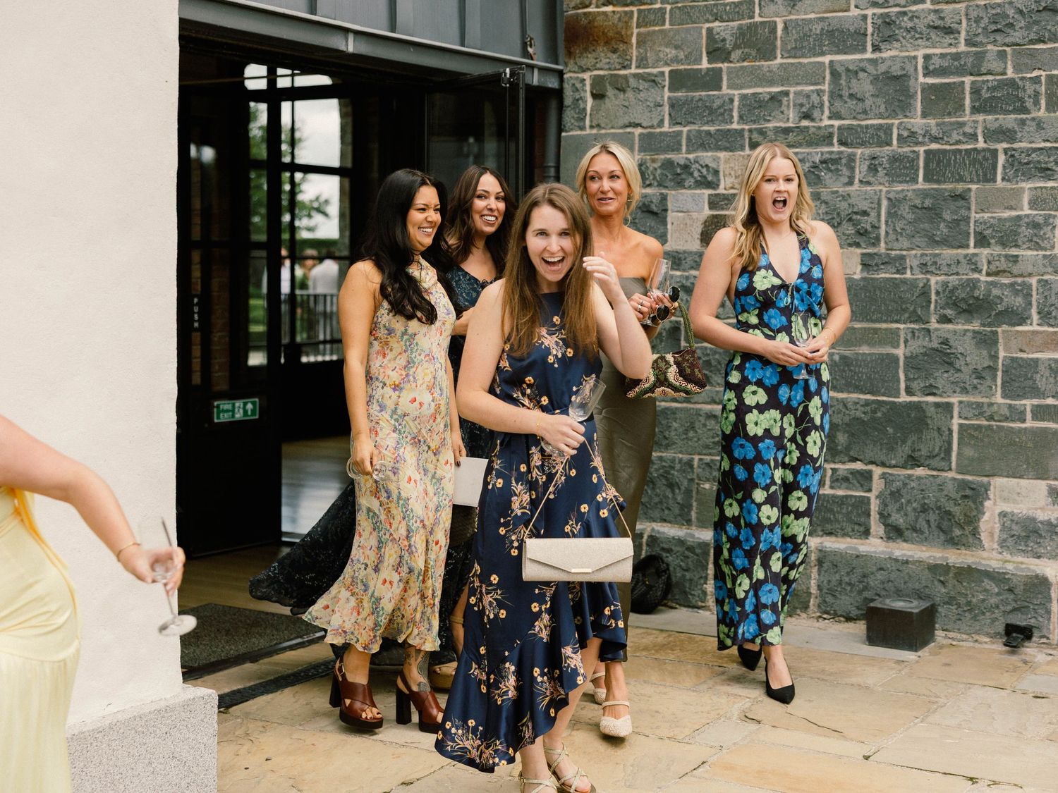 Wedding guests in floral dresses socialize outside a stone building.
