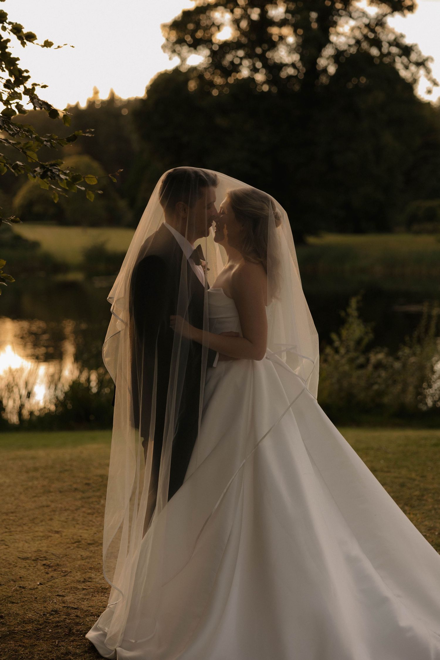 A romantic wedding couple embraces under a flowing veil at sunset by a lake.