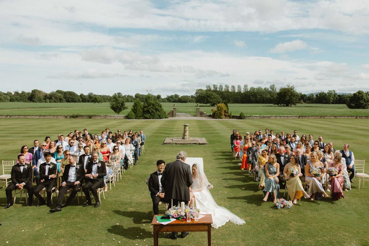 Outdoor wedding ceremony setup with rows of white chairs on a green lawn surrounded by trees.