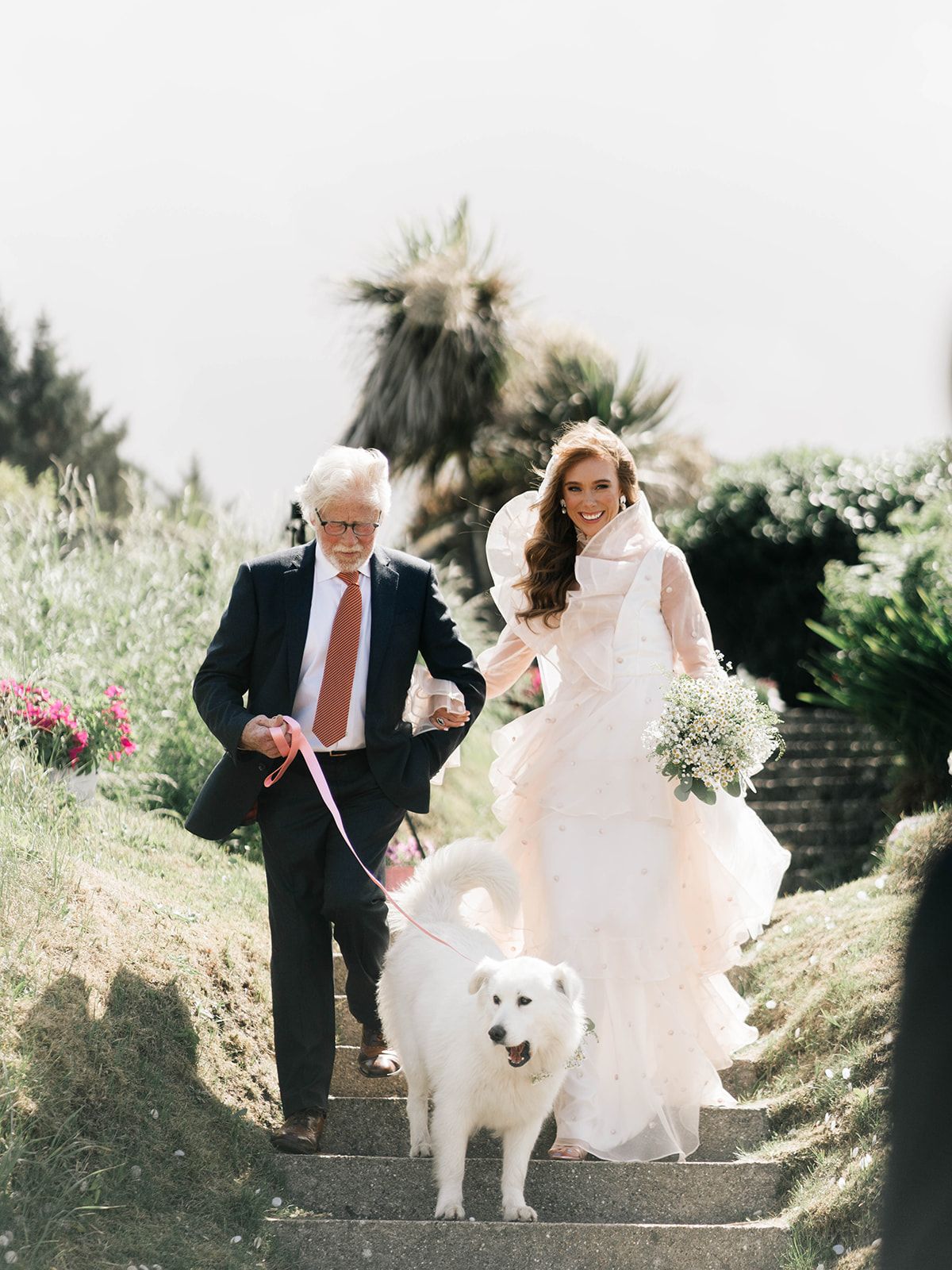 A bride in white dress walks down stone steps with an older man and fluffy white dog.