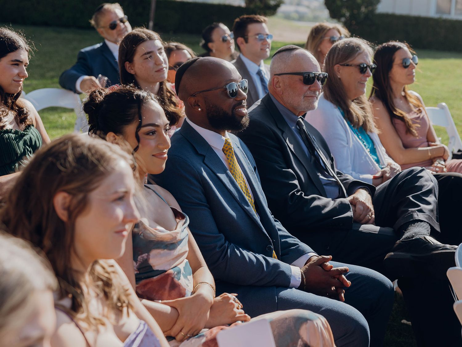 Wedding guests are seated outdoors in formal attire watching a ceremony on a sunny day.