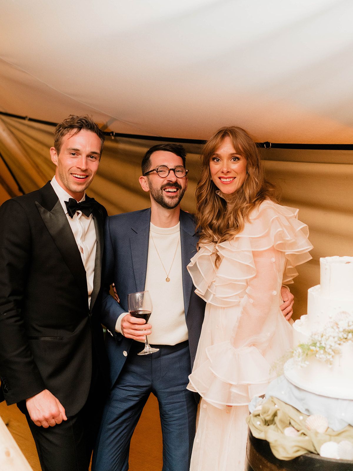 Wedding couple poses with guest by their white tiered cake at tent reception.