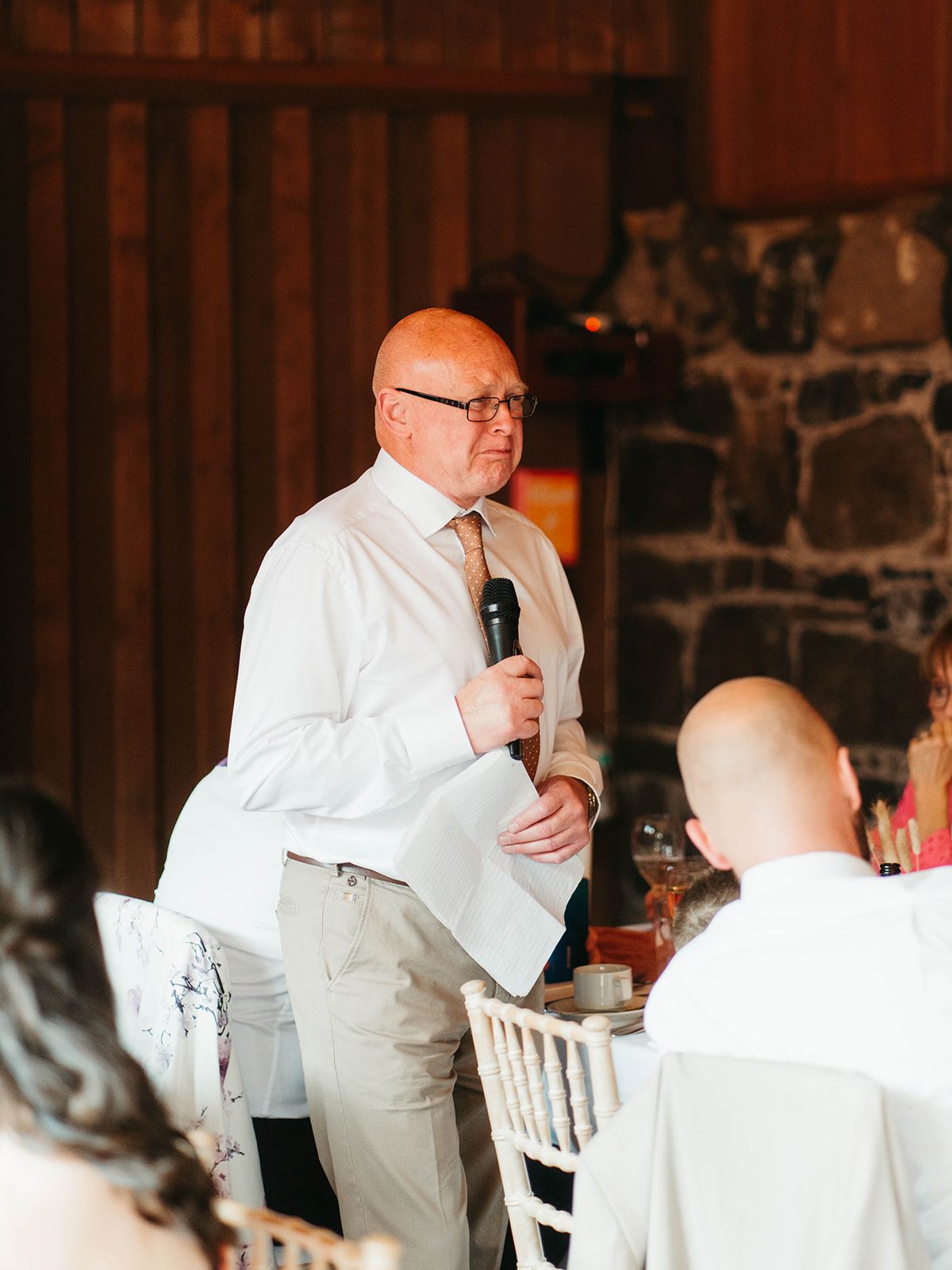 A person in a white shirt and khakis gives a speech with a microphone at an indoor event venue with wooden walls.