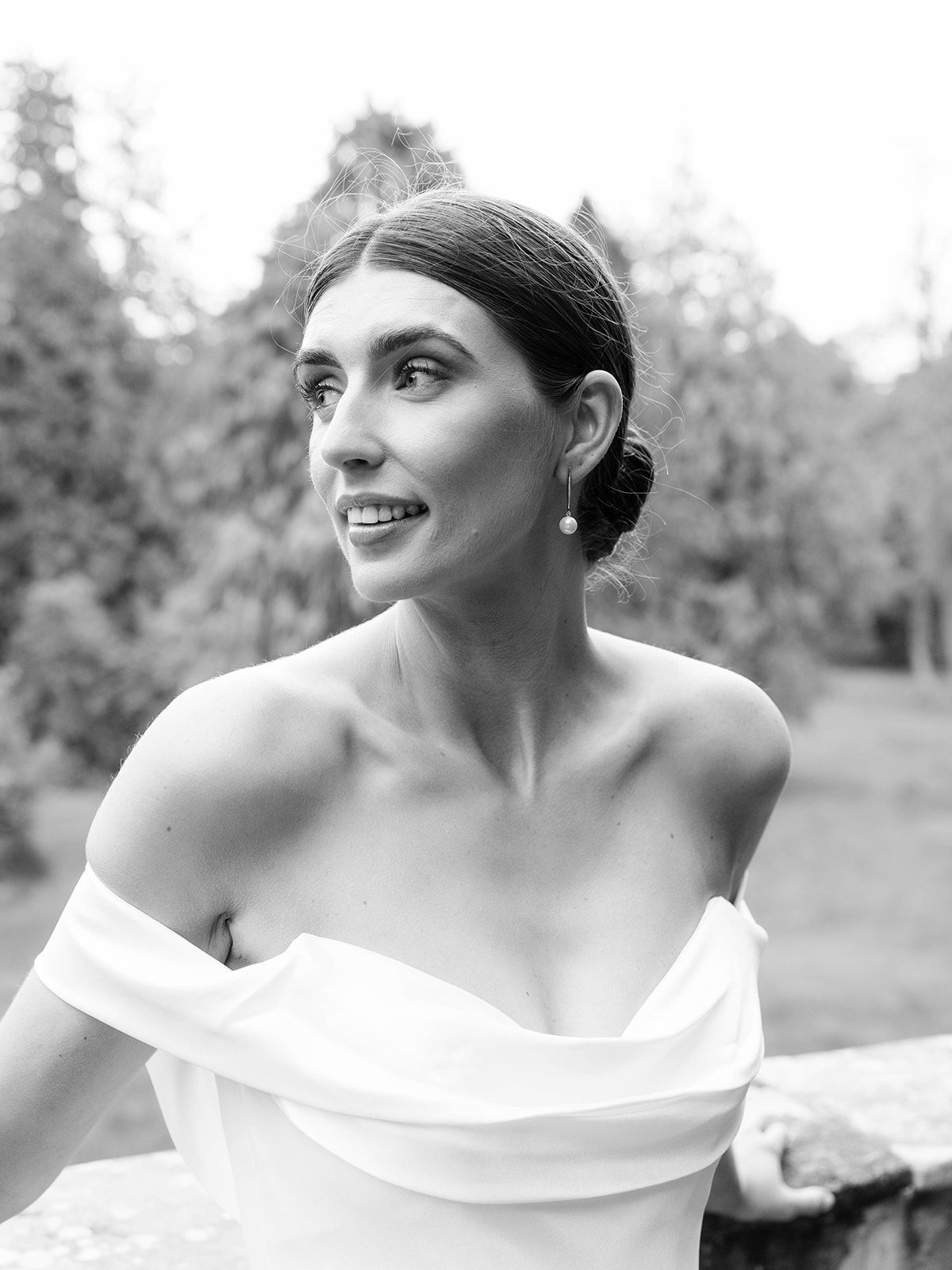 Elegant black and white bridal portrait showing woman in off-shoulder wedding dress looking to side with natural background.