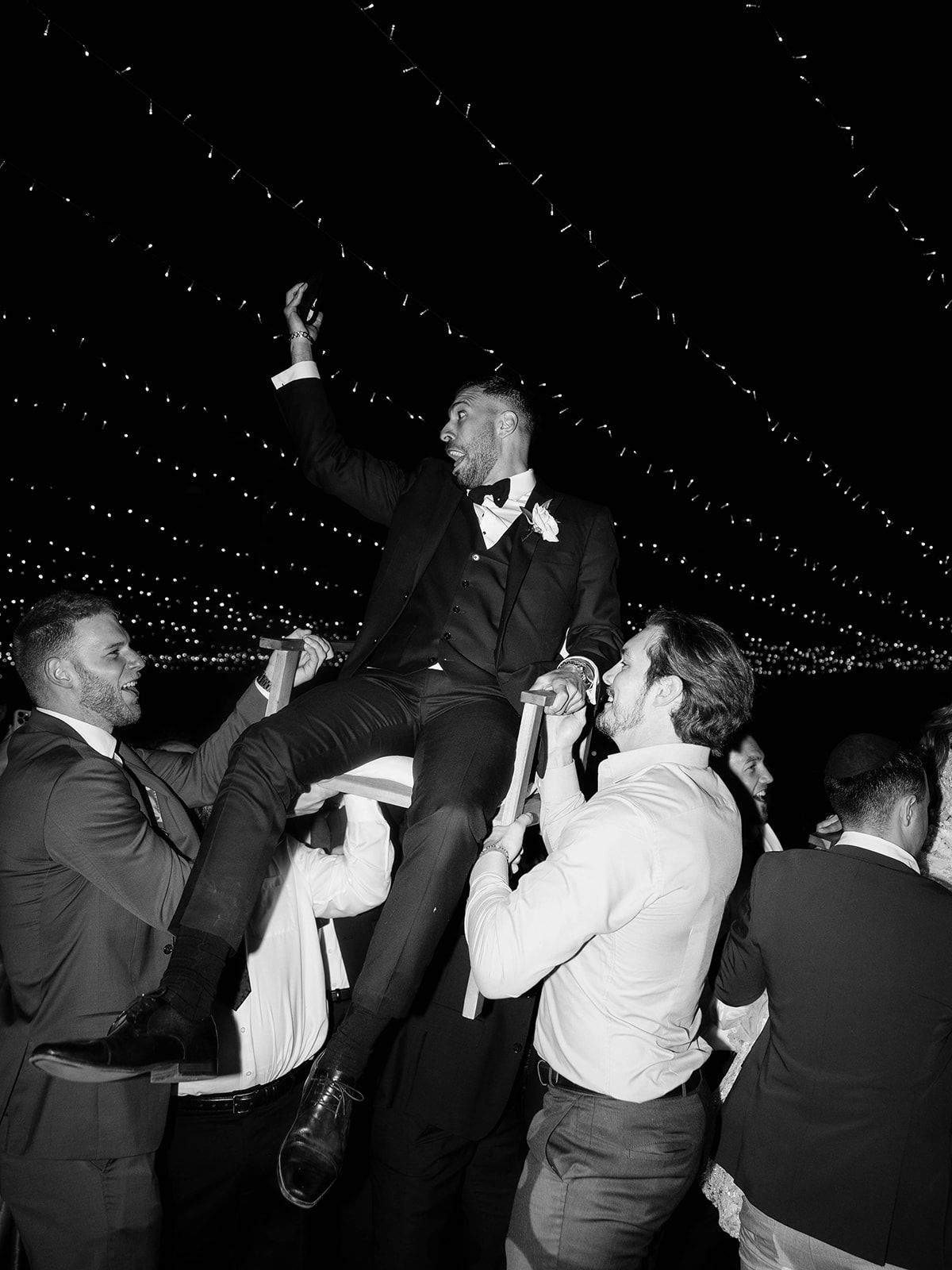 Wedding guests lift the celebrating groom in formal attire during an evening reception under twinkling string lights.