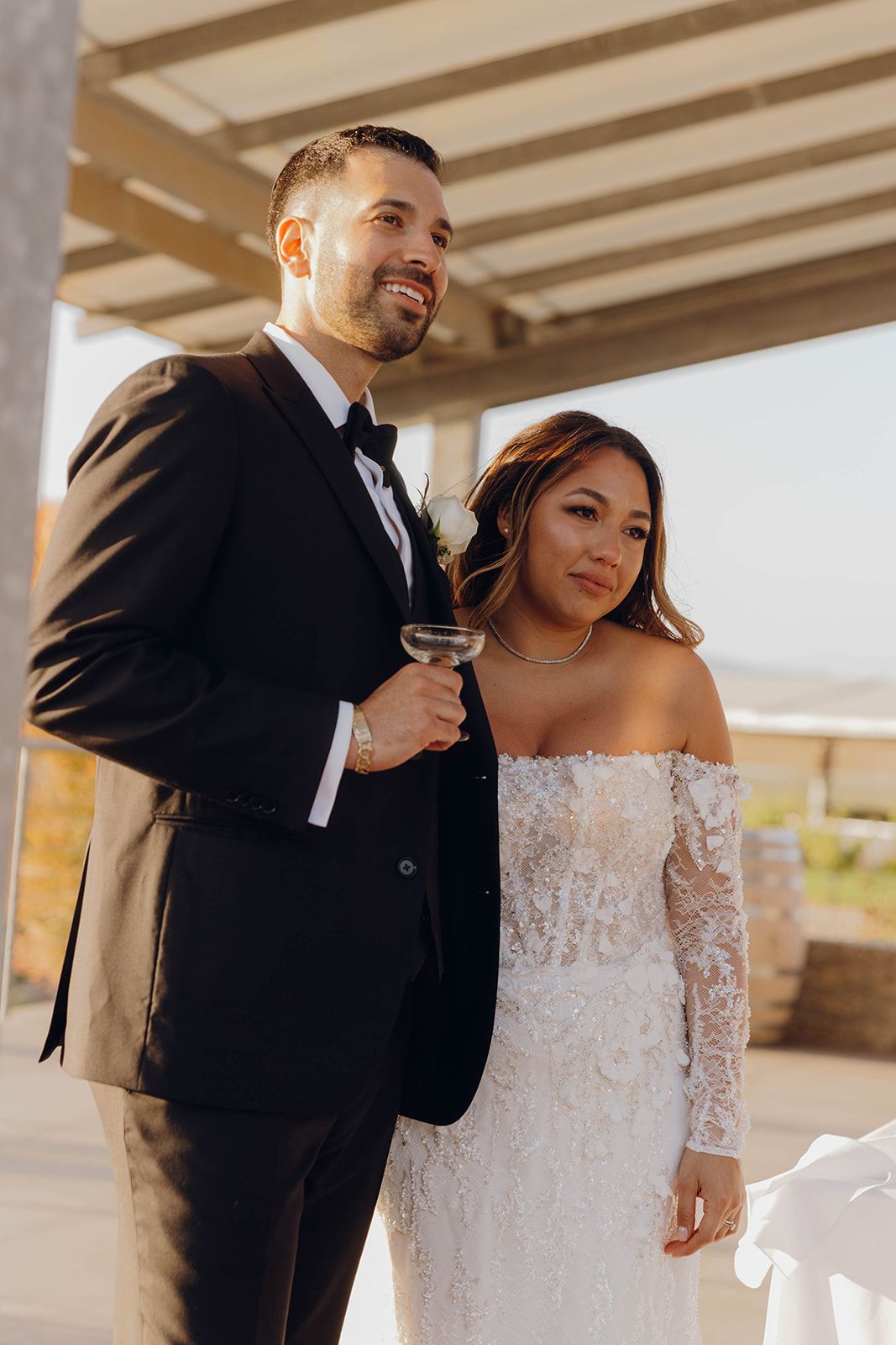 Elegantly dressed couple at outdoor wedding venue with bride wearing off-shoulder lace gown and groom in black tuxedo.
