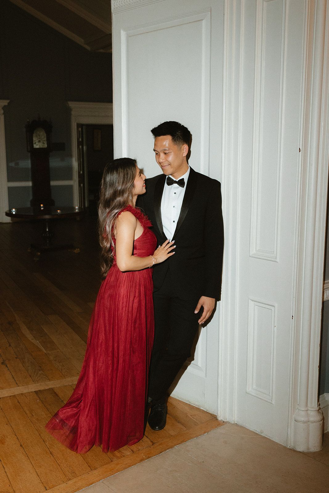 A couple in formal attire, with a woman in a red gown and a man in a black tuxedo, standing in an elegant interior.