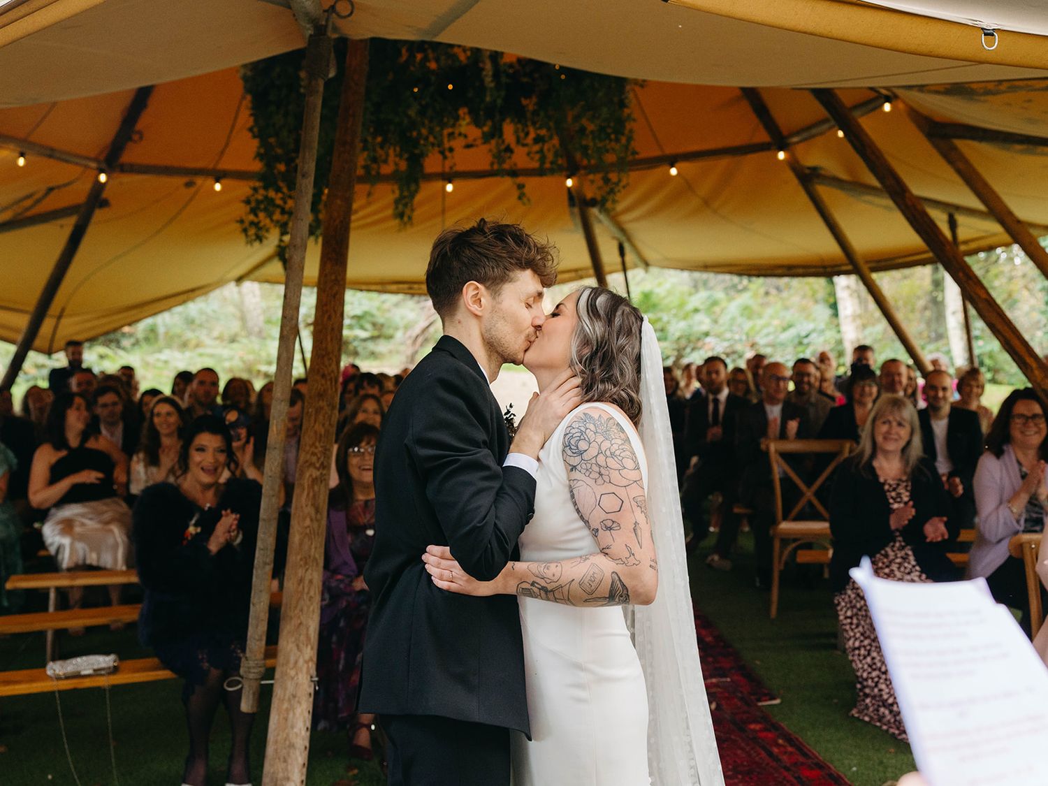 A couple shares their first kiss during their wedding ceremony under a rustic tent as guests look on.