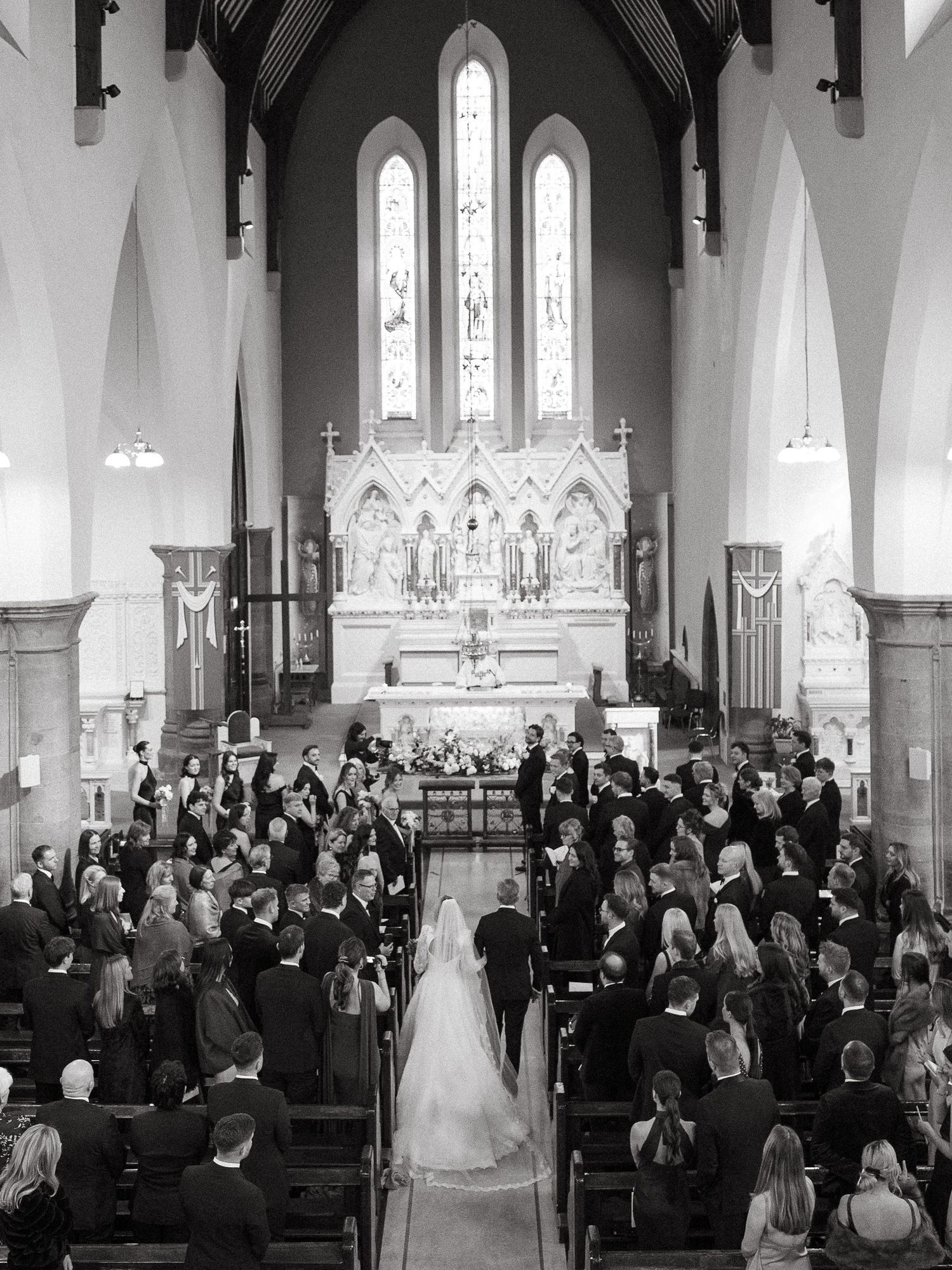 Black and white photo of a bride walking down the aisle in a grand church filled with wedding guests.
