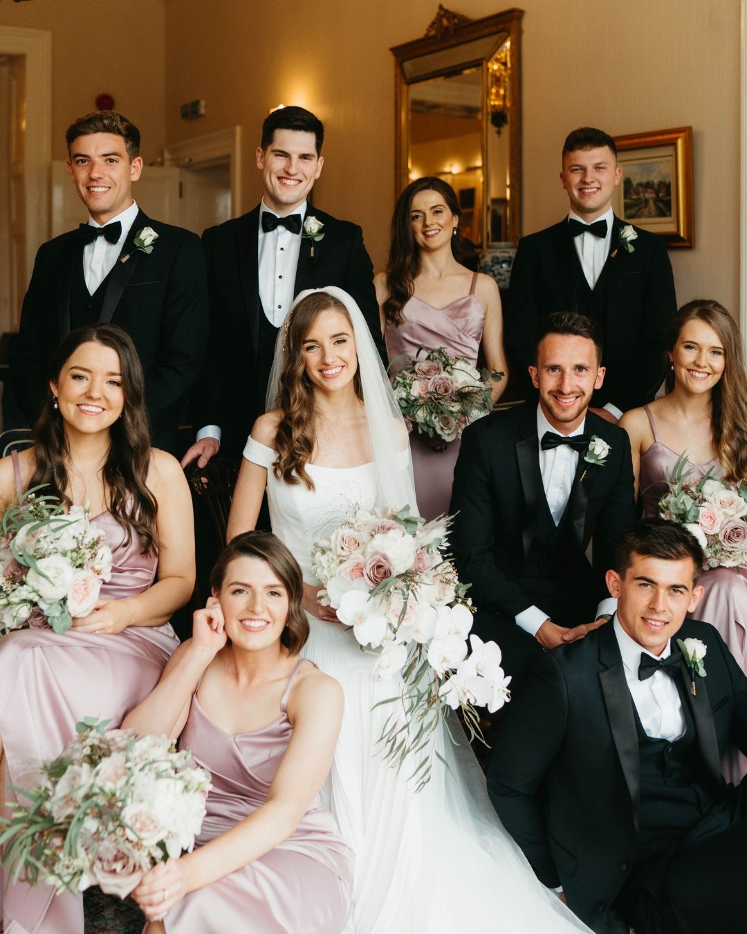 A wedding party in formal attire with bridesmaids in blush pink dresses and groomsmen in black tuxedos pose together.