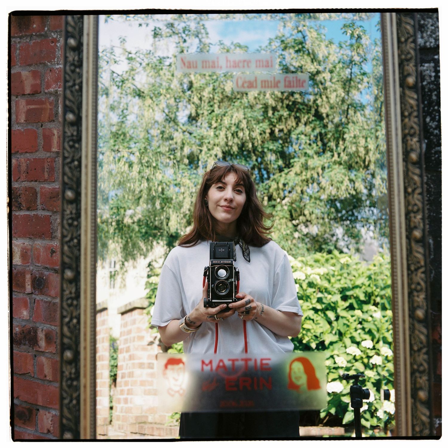 A young woman holds a vintage twin-lens reflex camera while standing in a sunlit doorway framed by brick walls.