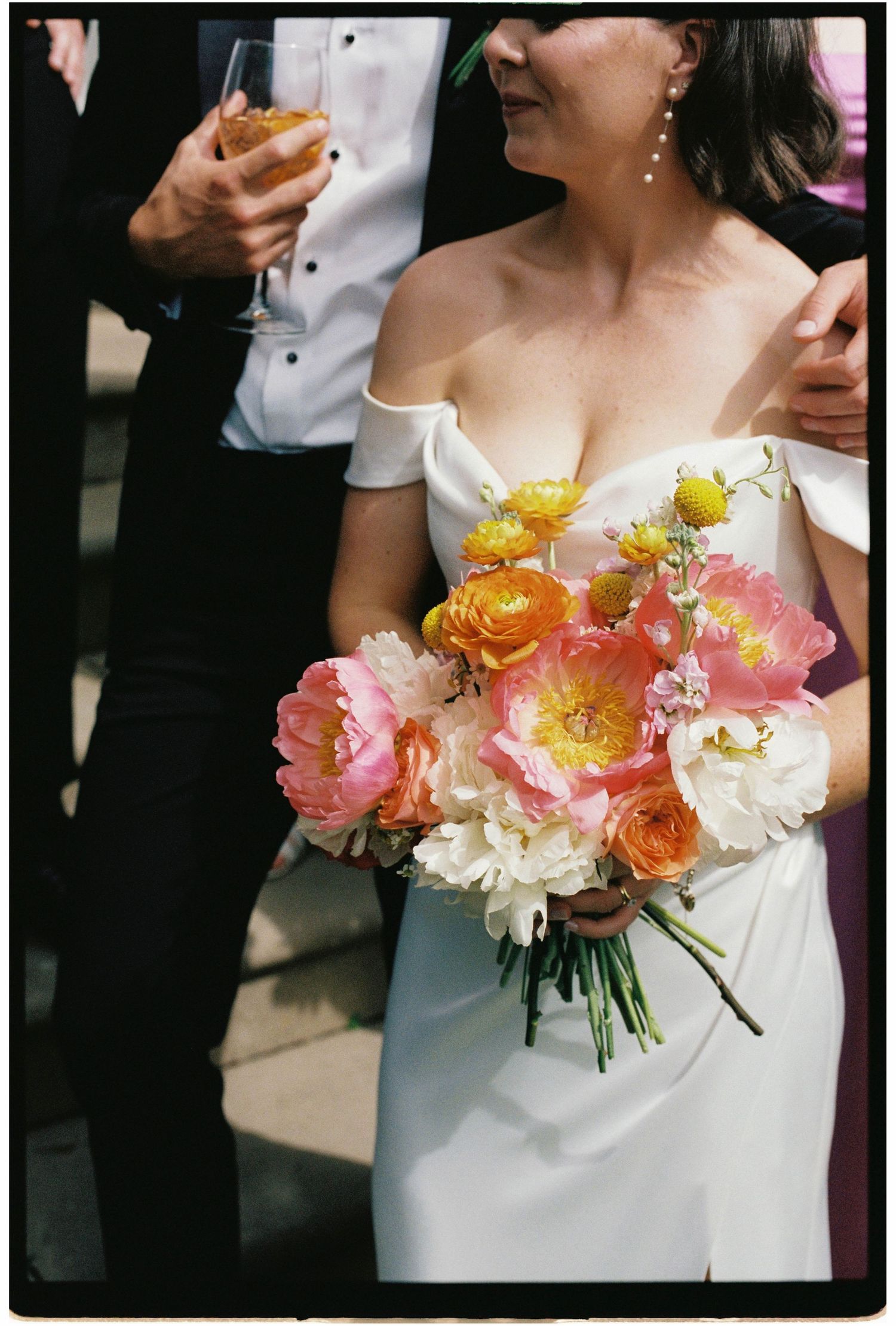 Bride in off-shoulder white gown holds a stunning bouquet of pink, orange, and yellow peonies at her wedding.