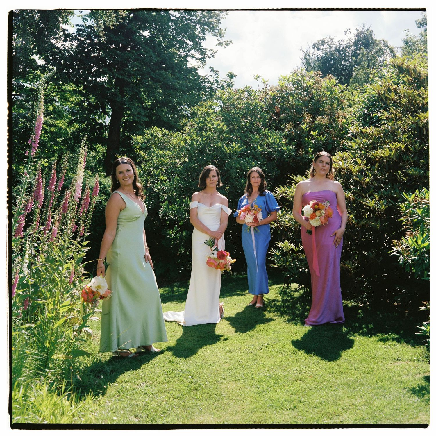 Four bridesmaids in colorful gowns holding bouquets stand on a sunny green lawn surrounded by lush garden foliage.