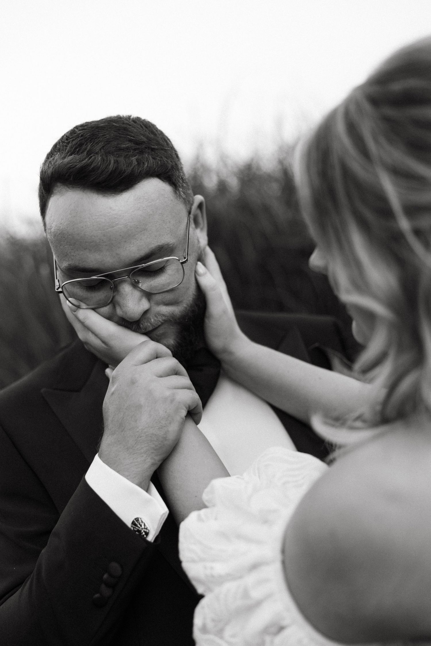 Black and white photo of emotional groom kissing brides hand during wedding ceremony outdoors.