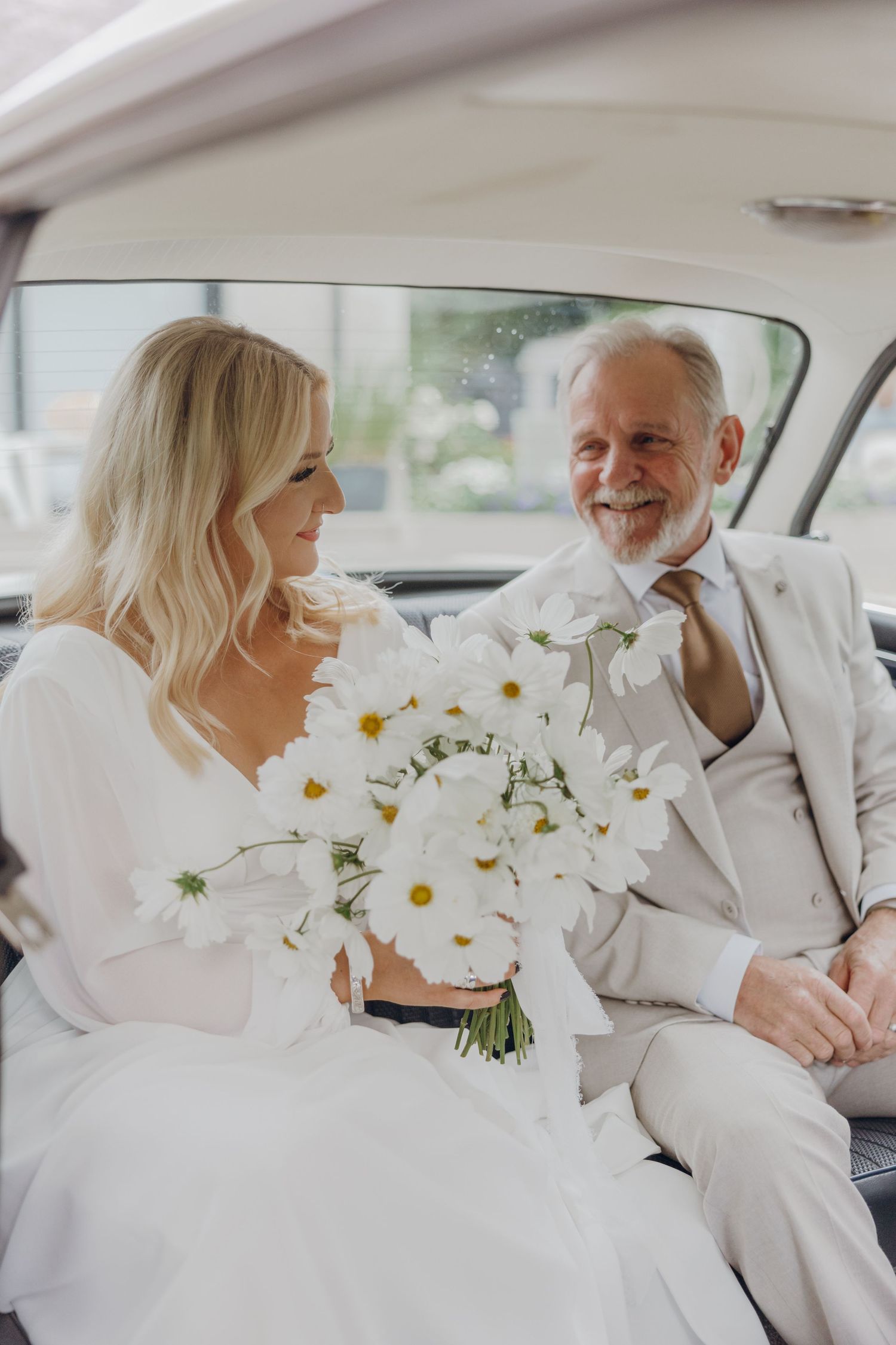 A couple dressed in white sits in the backseat of a car with a bouquet of white daisies between them.