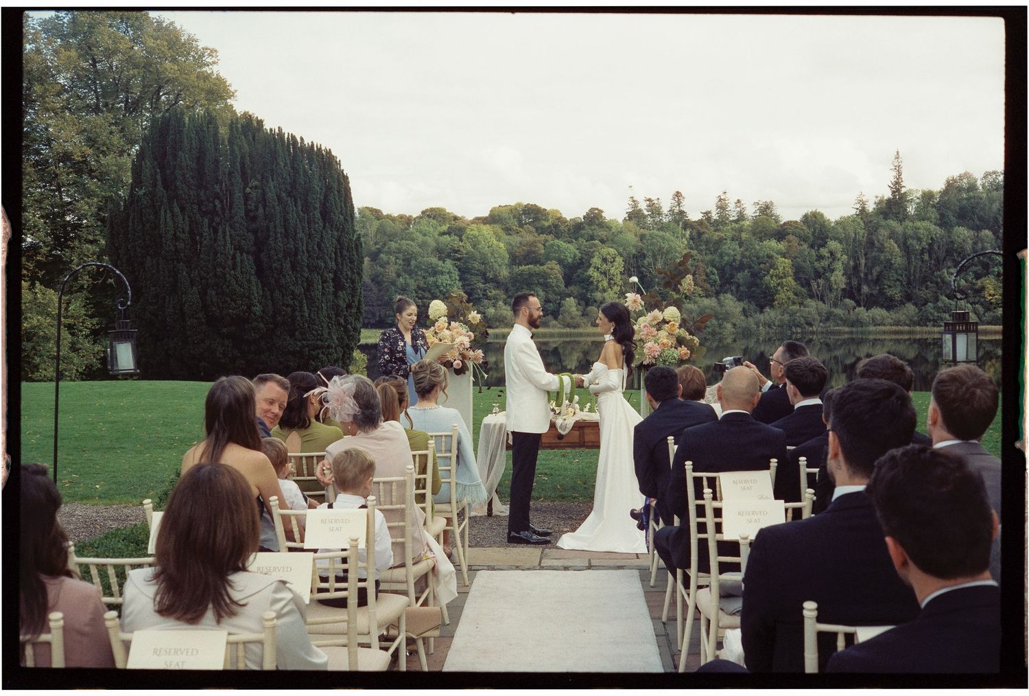 Wedding ceremony takes place in a beautiful outdoor garden setting with guests seated in white chairs on a lush green lawn.