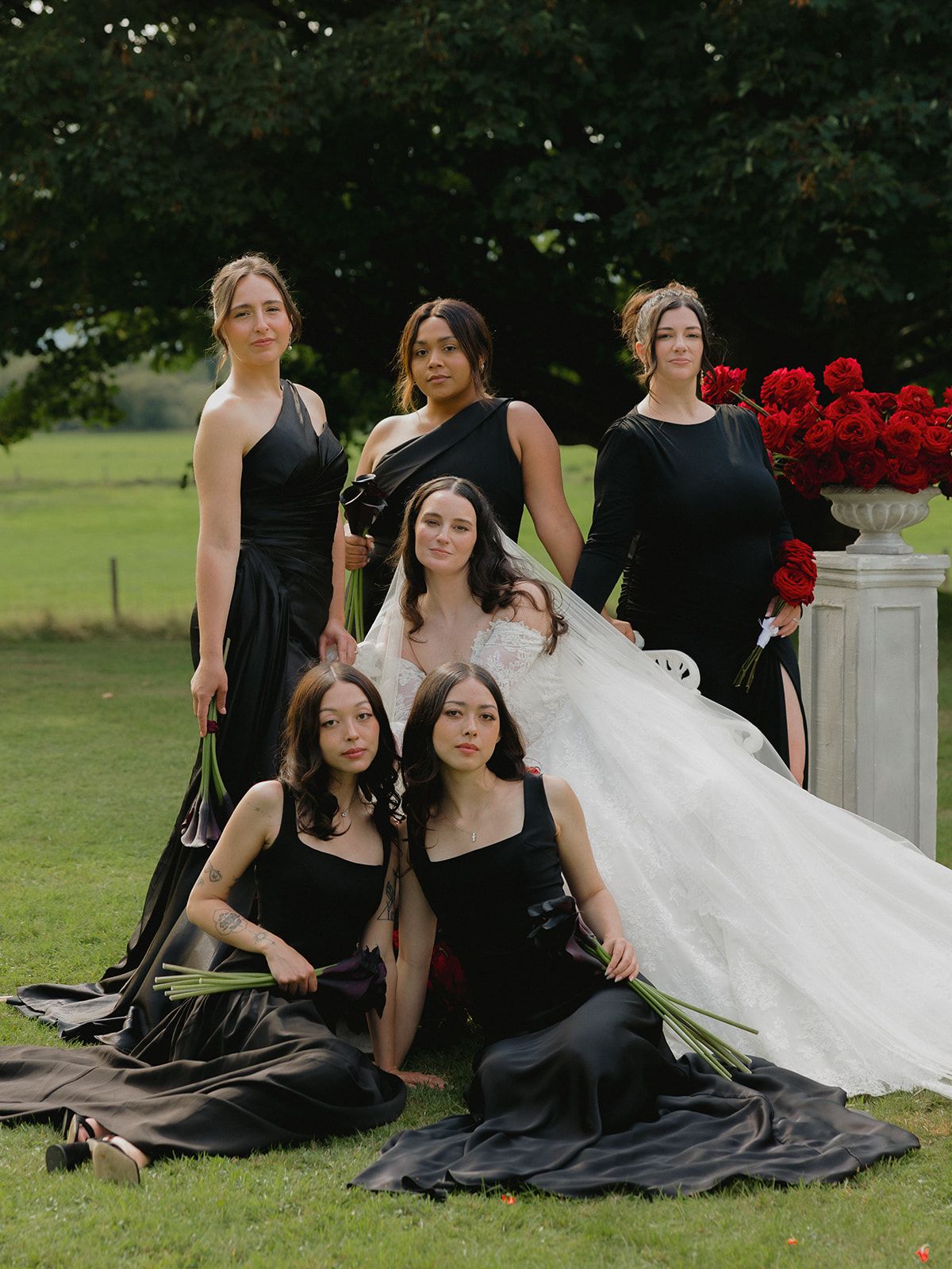 A bridal party in black dresses and a white wedding gown pose elegantly on a lawn with red floral decorations.