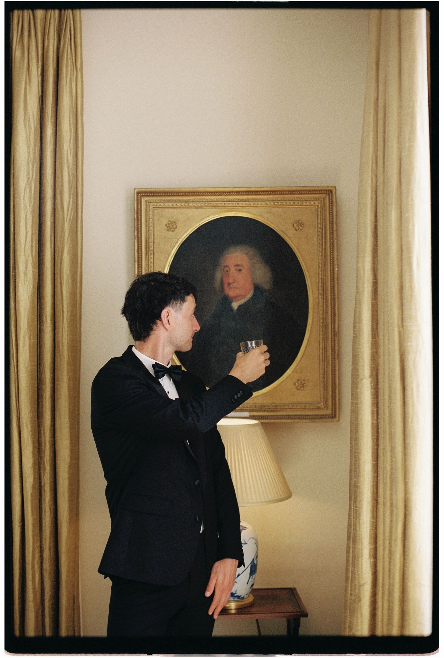 A groom in a tuxedo raises a glass toward a vintage oil portrait painting in an elegant interior setting.