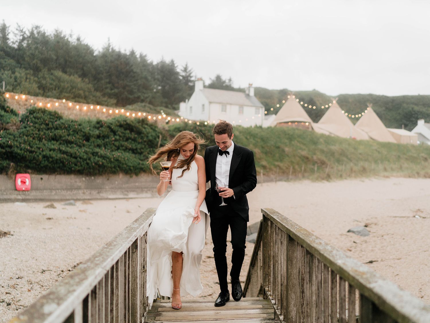 Couple walks along wooden boardwalk with beach tents visible in background.