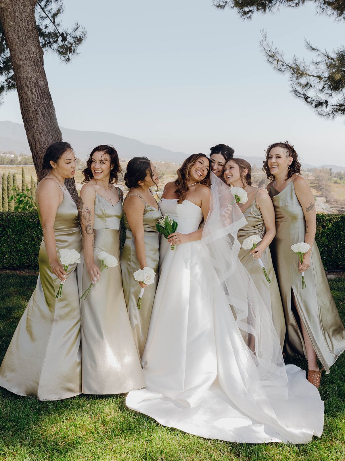 Bridesmaids in champagne silk dresses gather around a bride in a flowing white gown on a scenic hilltop.