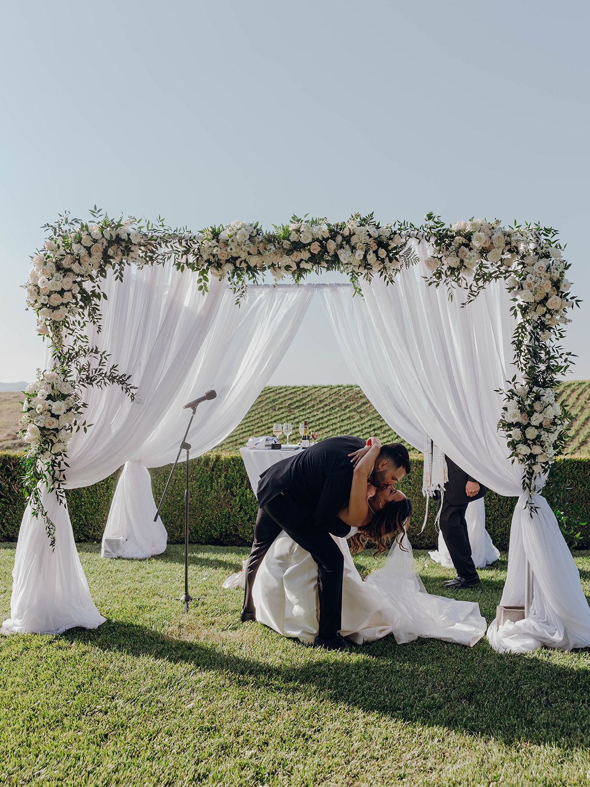 A beautiful outdoor wedding ceremony setup with a white draped arch decorated with floral arrangements on a grassy lawn.