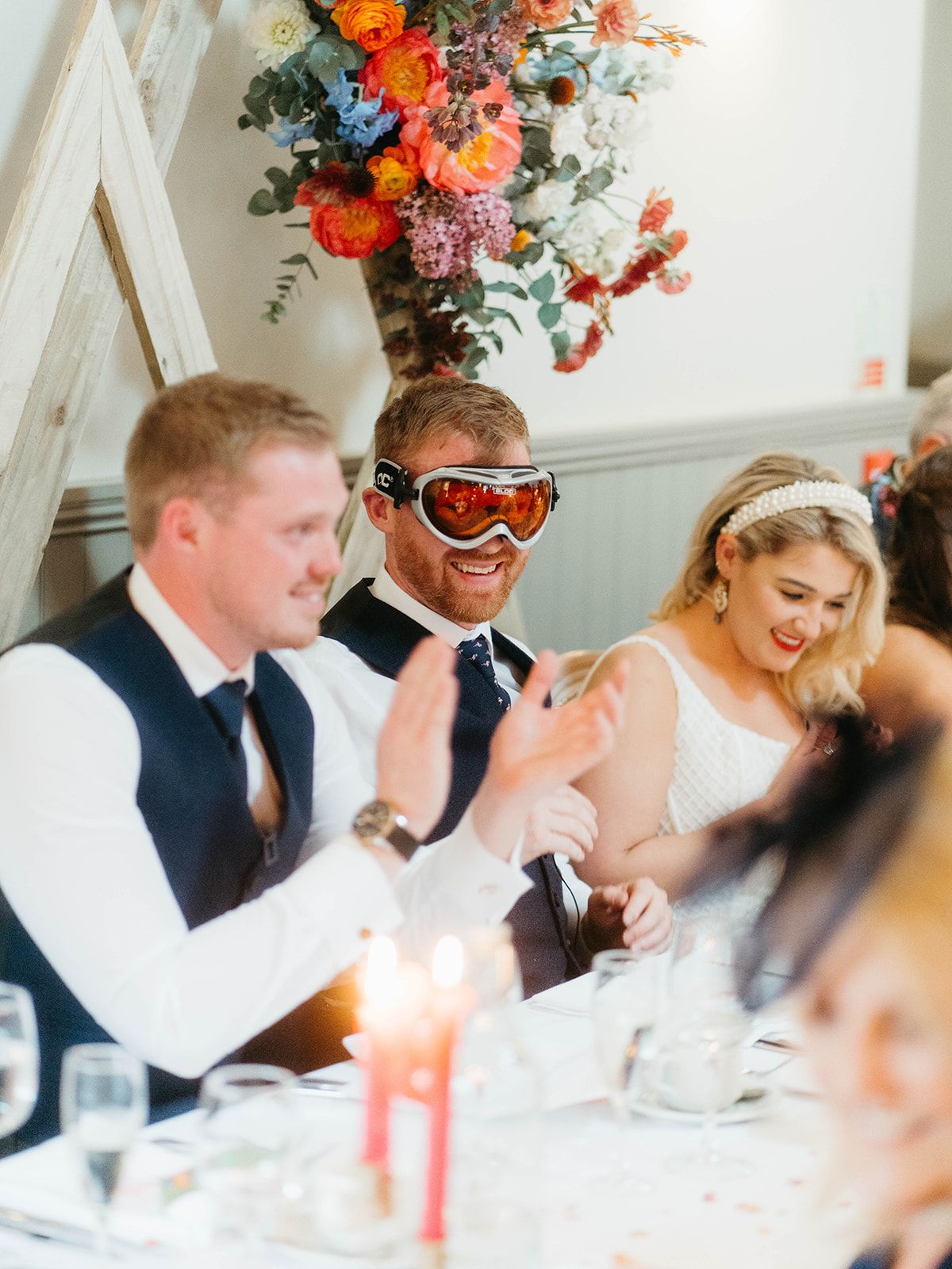 Wedding reception guests share a joyful moment at a dinner table beneath vibrant floral arrangements.