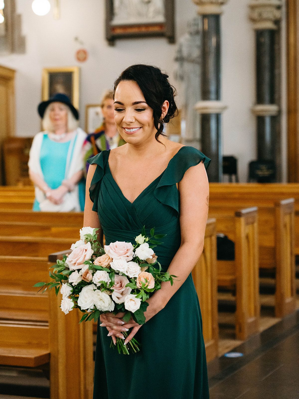 Bridesmaid in emerald green dress holding pink and white floral bouquet stands in church aisle during ceremony.