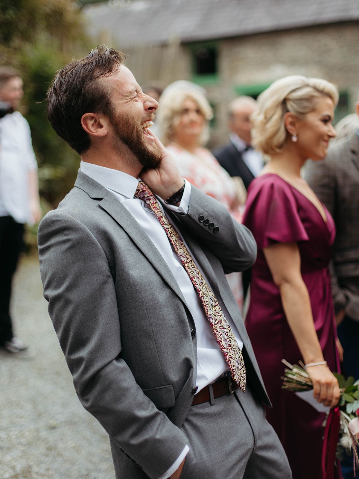 Wedding guest in grey suit and patterned tie laughs heartily at outdoor celebration while others gather in background.