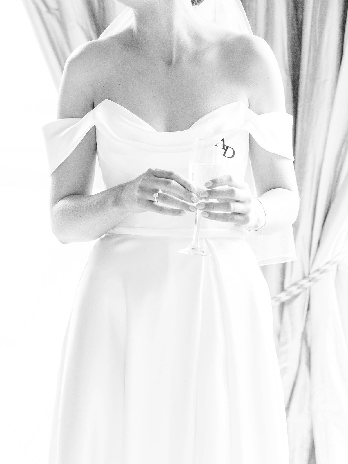Bride in classic white off-shoulder gown holds a delicate crystal glass against soft fabric backdrop.