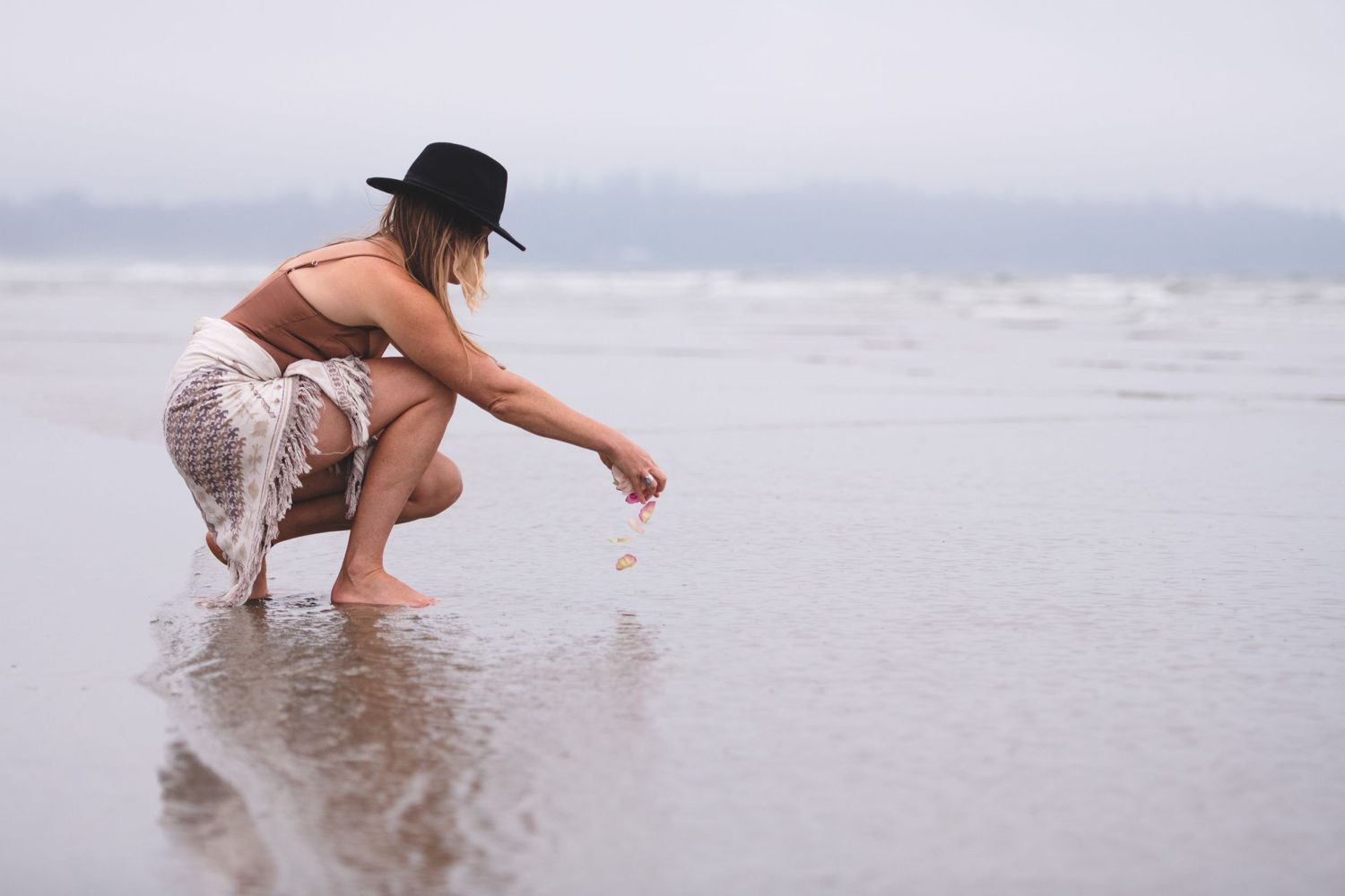 Person in black hat crouches down to touch ocean water at beach.