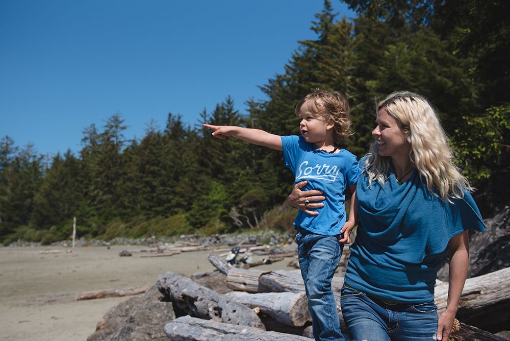 Two people in blue shirts sit on driftwood at a beach with evergreen trees in the background while pointing into distance.