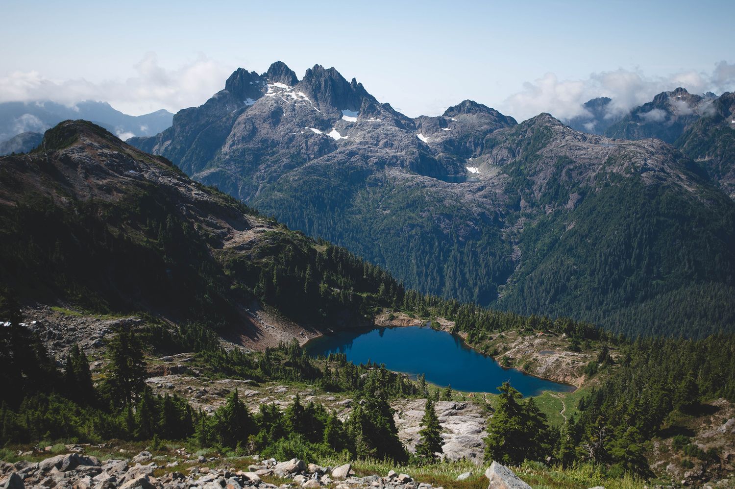 Aerial view of a turquoise alpine lake surrounded by dramatic mountain peaks and evergreen forest.