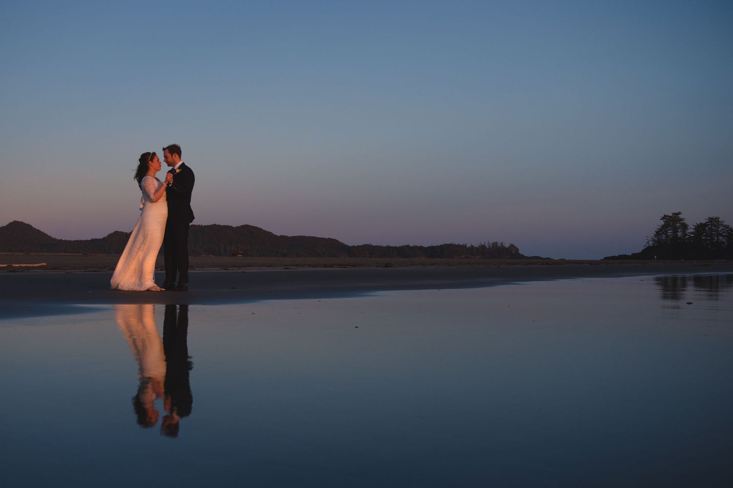A couple's reflection mirrors in still water at dusk with mountains in background.