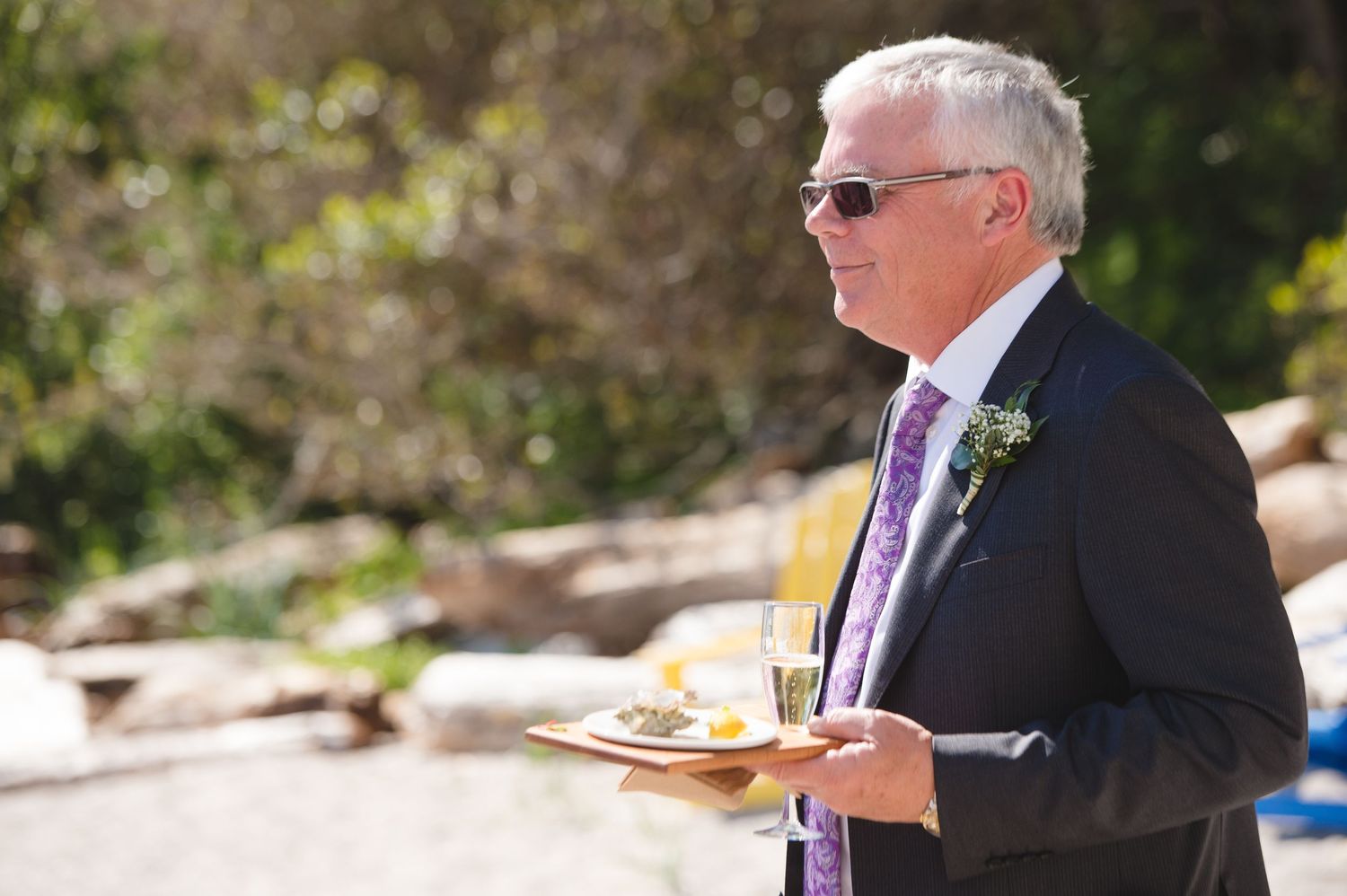 Wedding guests seated in formal attire on a rocky beach with evergreen trees in background.