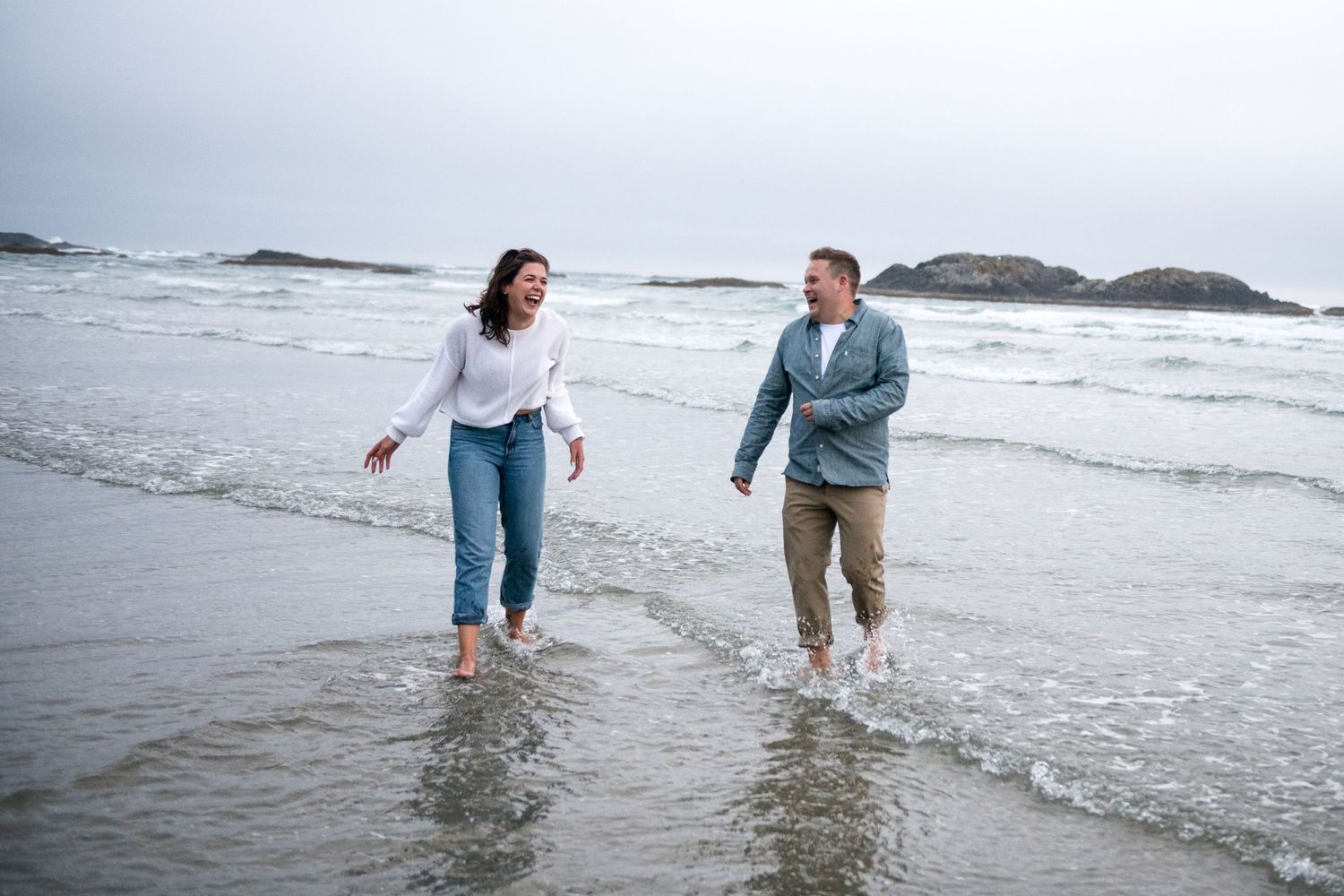 Two people splash and play in the shallow ocean waves on an overcast day.