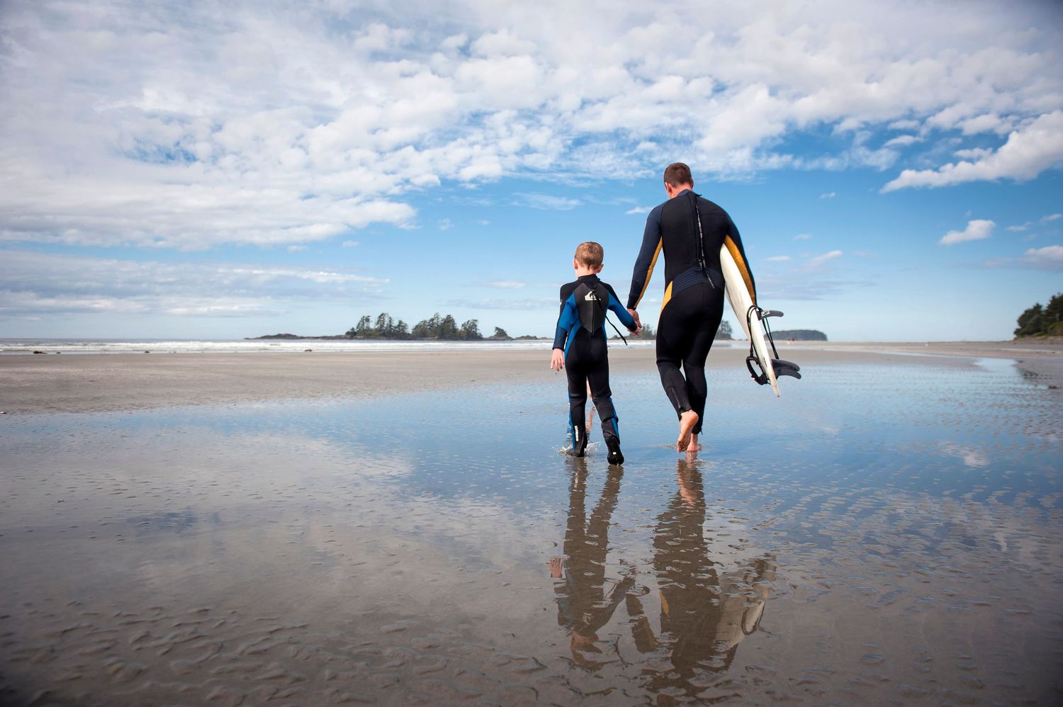 Father and son surfers walking along a Tofino beach at low tide with their surfboards and reflections in the wet sand.