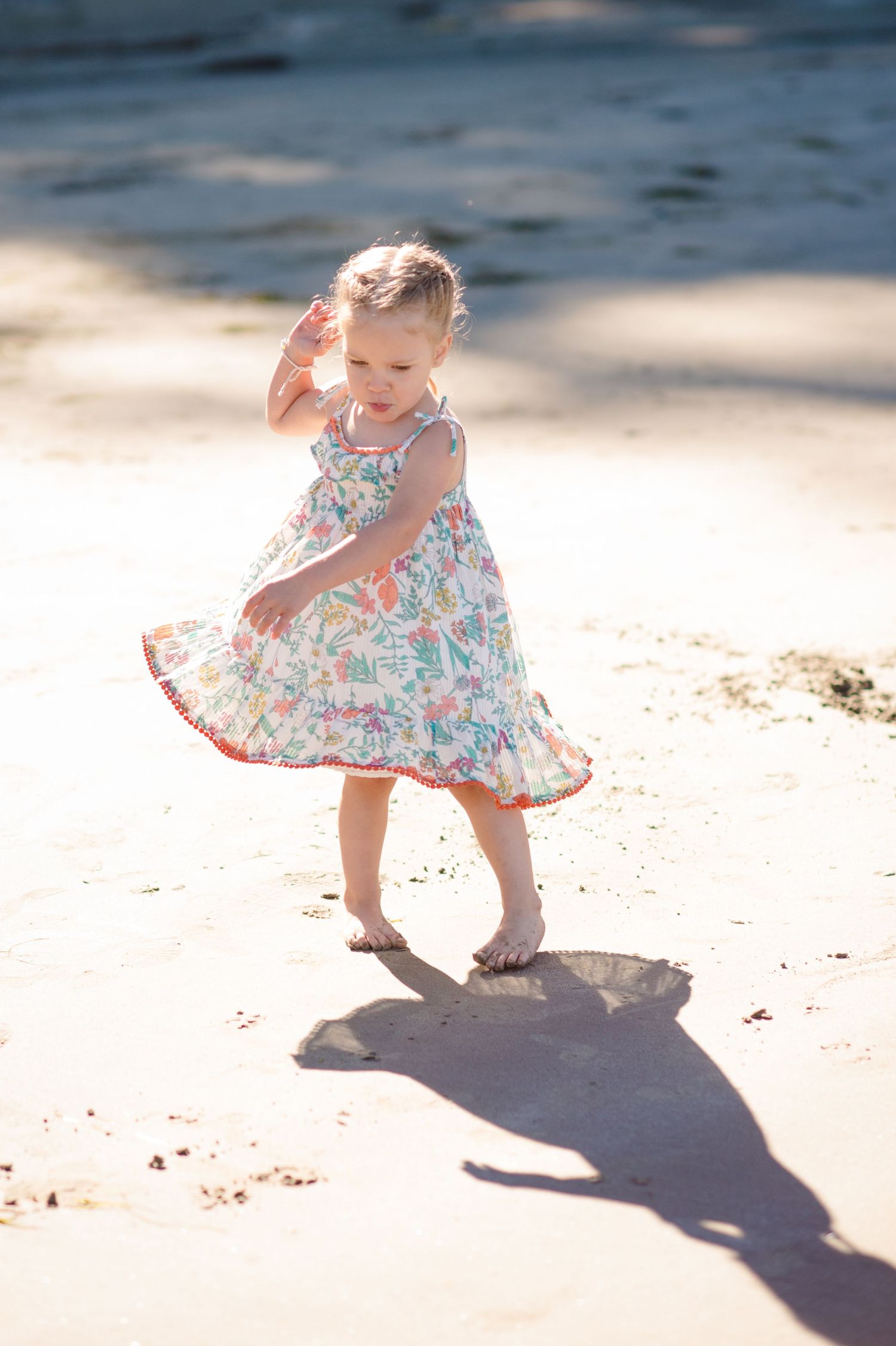Child in floral dress casting long shadows while playing on sandy beach during golden hour.