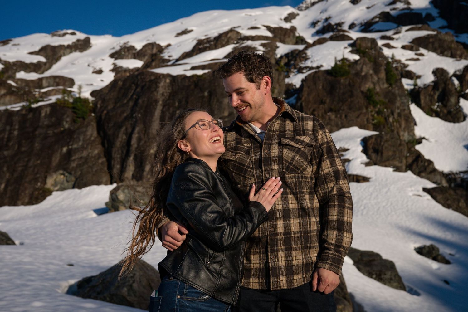 Happy couple laughing together on a snowy mountain landscape with rocky cliffs and patches of snow behind them.