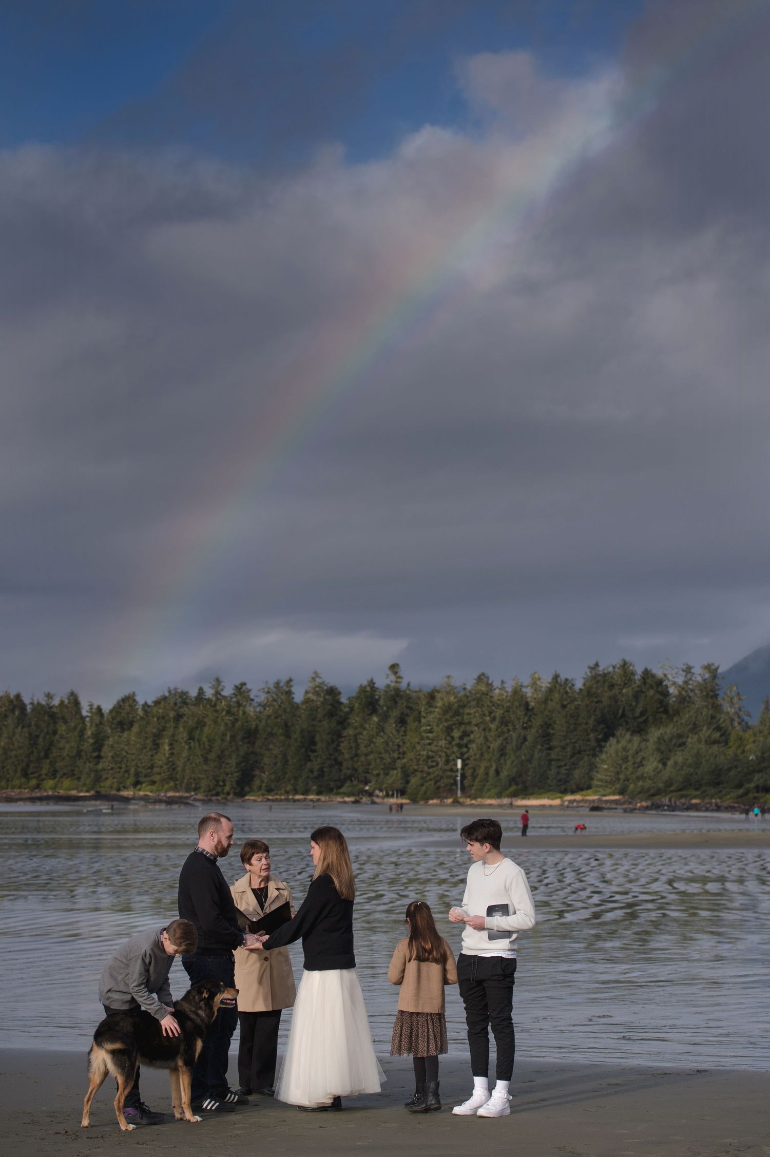 A rainbow arches over a coastal wedding ceremony setup with mountains in the background.