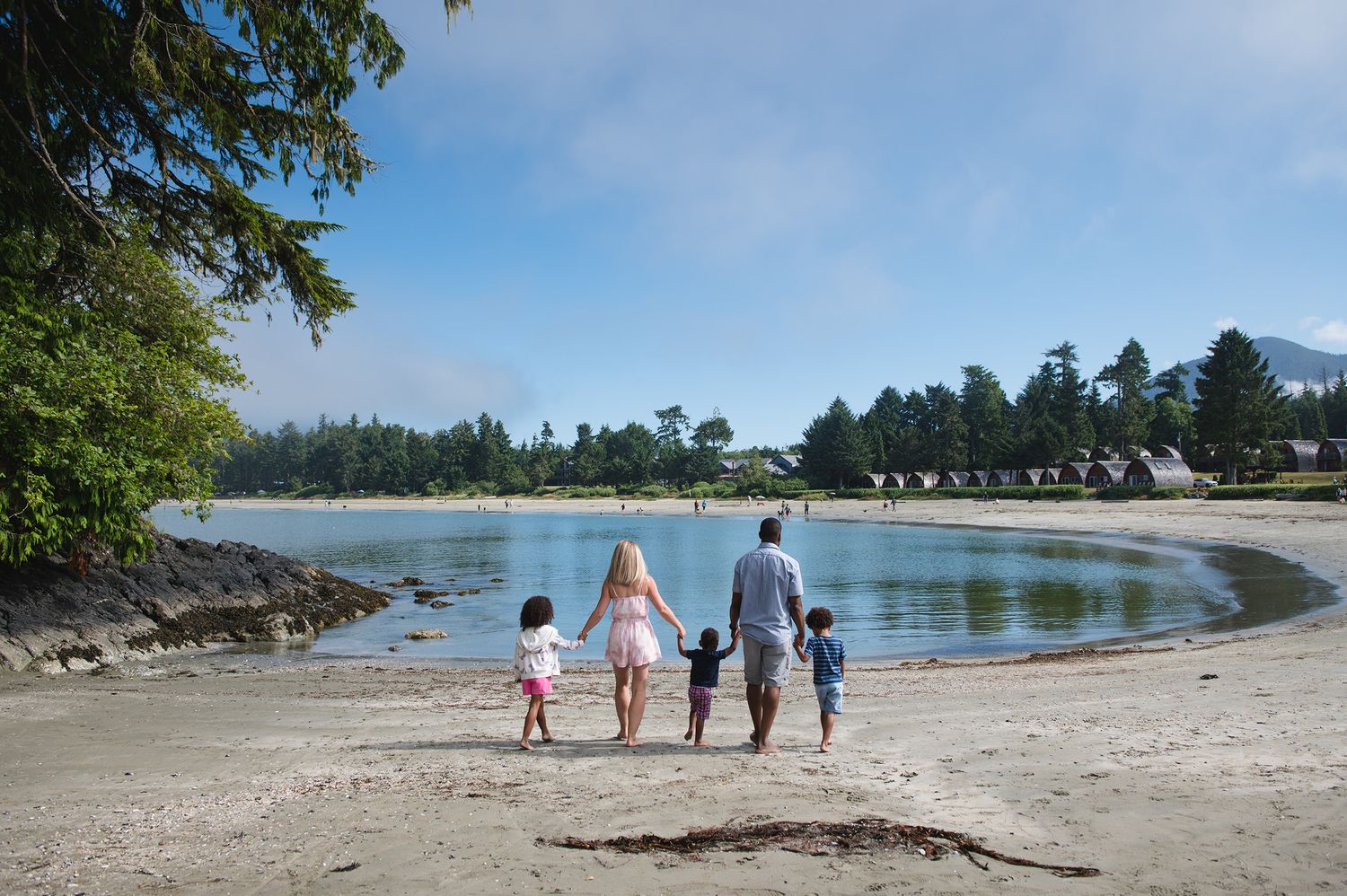 Scenic wide view of curved beach coastline with evergreen trees and people walking in distance.