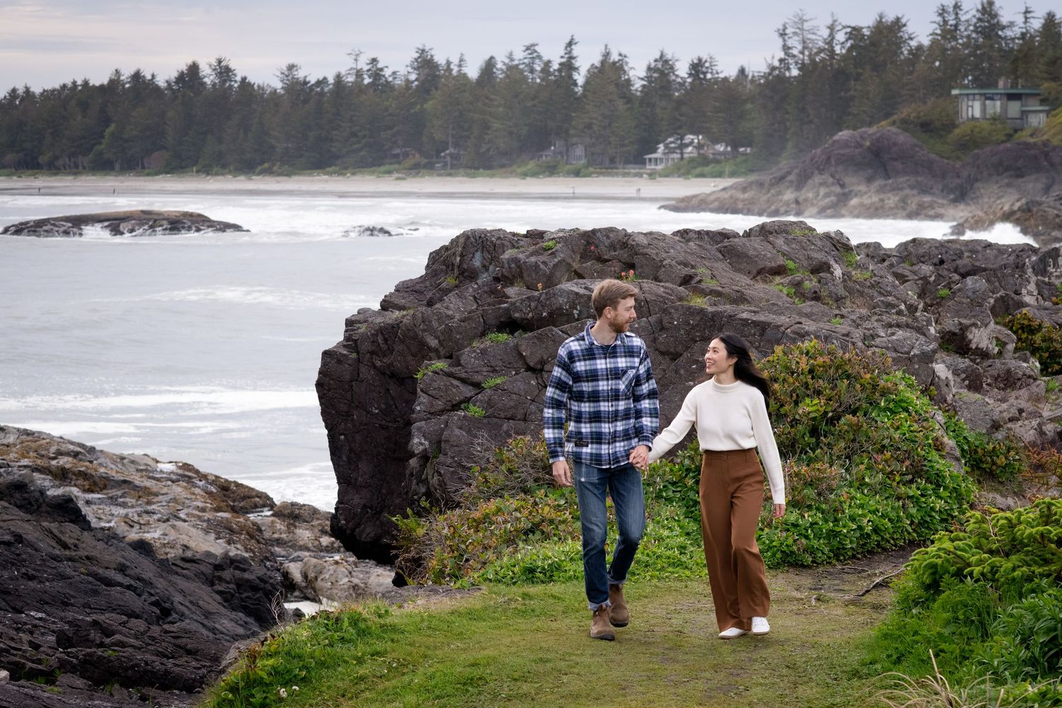 People exploring a dramatic rocky shoreline with waves crashing against volcanic rocks and green vegetation.