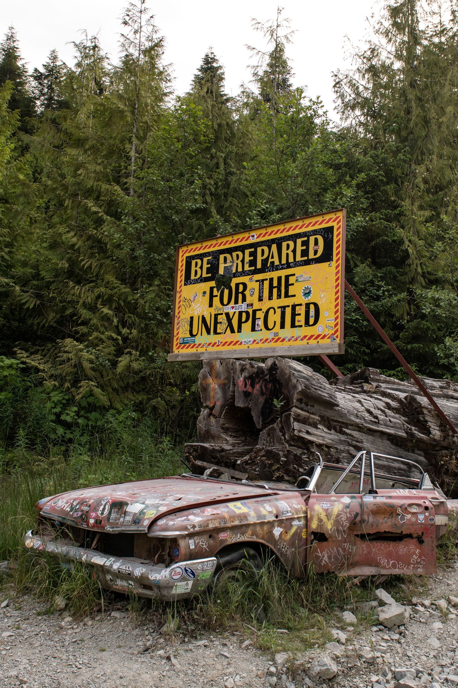 Rusty abandoned car beneath warning sign reading 'Be Prepared for the Unexpected' against forest backdrop.