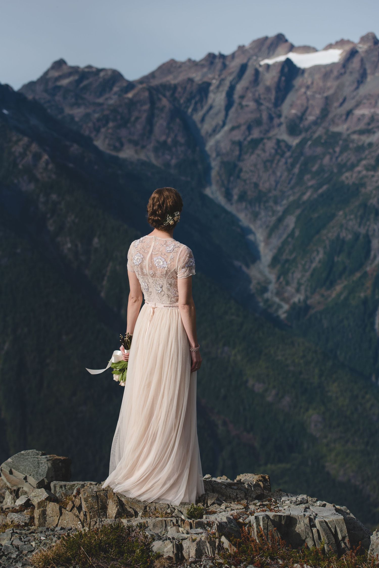 Person in a flowing dress stands at mountain overlook with dramatic peaks in the distance.